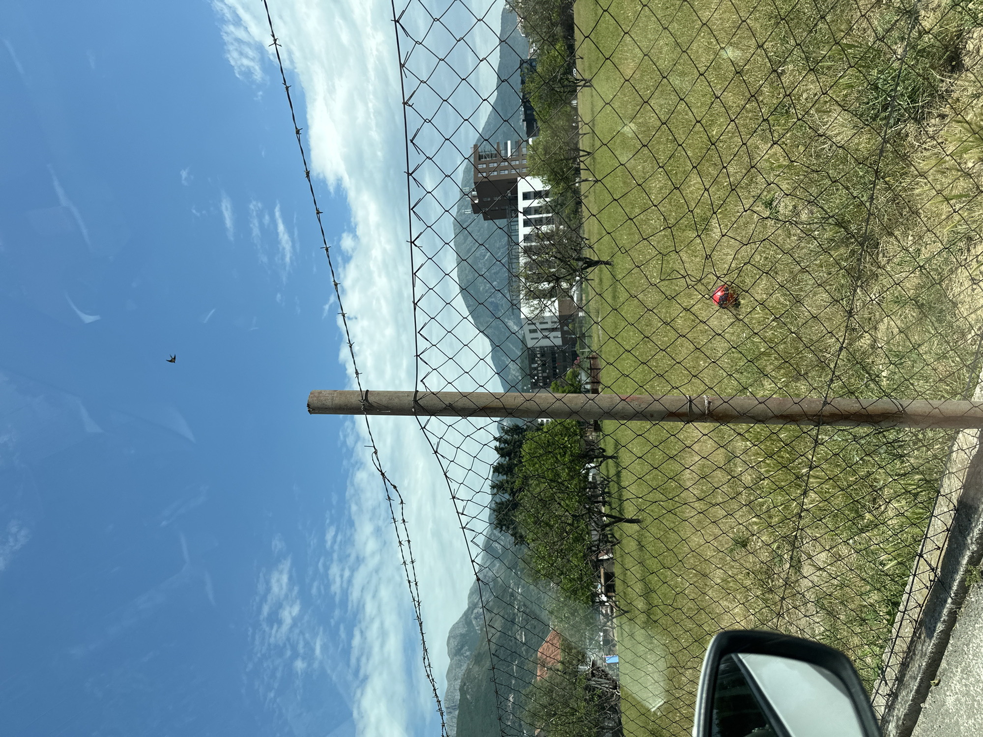The image, rotated for easier viewing, shows a chain-link fence with a metal pole support and barbed wire at the top. The fence encloses a grassy area with patches of dirt and scattered vegetation. Beyond the fence, a building with white and brown exterior elements is visible against a backdrop of mountains. The sky is blue with scattered white clouds. A bird is flying in the sky. Part of a car side mirror is visible in the lower right corner of the image. A red object rests on the ground within the fenced area. The location appears to be Bar, Montenegro, based on the architecture and mountainous terrain.