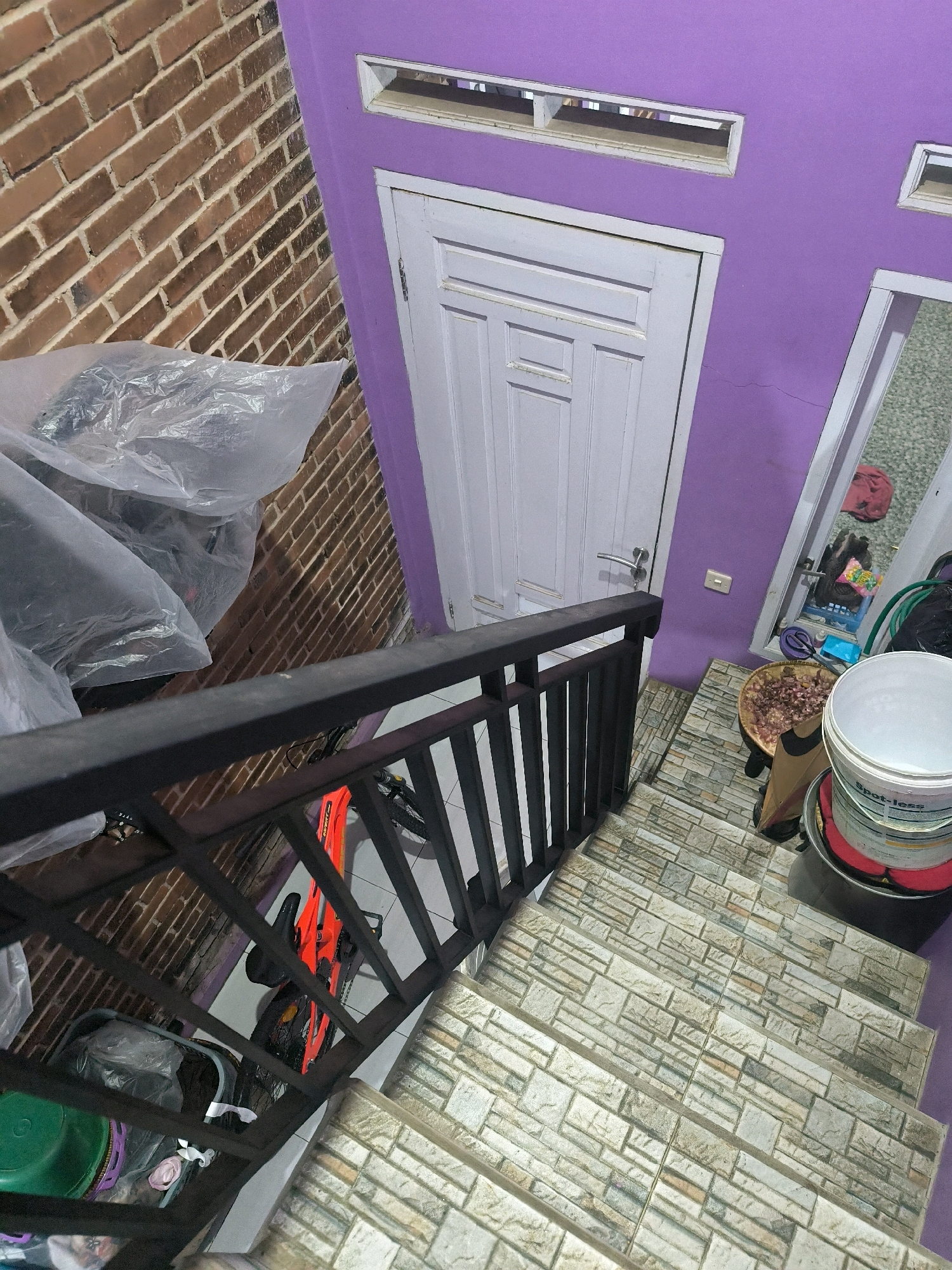 This media file depicts an outdoor stairway leading down from a balcony or upper level. The scene is an outdoor setting, likely residential, given the brick wall and purple painted exterior wall. The stairway is covered with stone-like tiles and has a black metal railing.
At the top of the stairs, there is a white door with a metallic handle. To the right of the door, a reflection in a mirror shows various items including laundry, a green hose, and a white bucket labeled "Spot-less". There is also a woven basket filled with chopped onions.
On the left side of the stairs, a red bicycle is visible, partially covered by clear plastic sheeting. A brick wall is present on the left, and the purple painted wall continues to the right. Two rectangular windows with louvers are mounted high on the purple wall.
The overall impression is one of a functional, perhaps slightly cluttered, residential area. The lighting suggests it is daytime, though no specific time can be determined. The location context is Cilengkrang, Indonesia. There is no visible text other than the "Spot-less" label on the bucket.