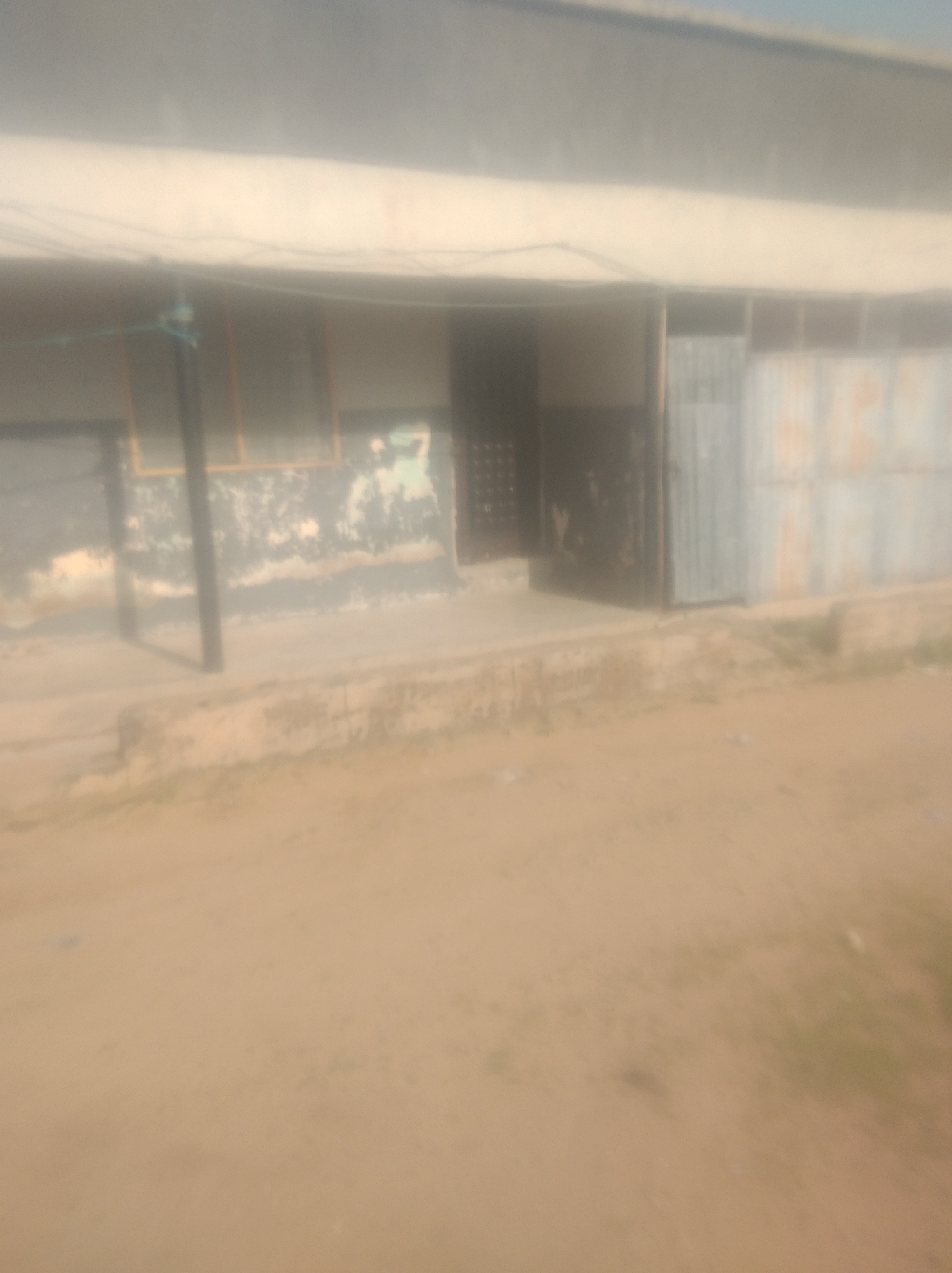 The media file depicts an outdoor scene, likely in Jalingo, Nigeria. The primary subject is a weathered building with a light-colored, possibly concrete, facade. The building shows signs of age, with peeling paint and some discoloration. It features several openings, including a large metal door or gate on the right, appearing to be corrugated metal. To the left of this, there's another dark opening, possibly a doorway, with some texture suggesting a grate or patterned surface. Further to the left, there are window-like structures with orange frames, partially obscured. A dark, vertical pole stands in front of the building, possibly a support or part of a fence. Electrical wires or cables are visible strung across the upper portion of the building's facade. The ground in front of the building is a dusty, light brown earth, with a raised concrete or stone curb running along the base of the structure. The overall impression is one of a utilitarian building in an unpaved area, possibly a workshop, storage facility, or older commercial space. The lighting suggests it is daytime, and there are no obvious cues about the weather, though the dusty ground might imply dry conditions. No people are visible in the frame, and no text is discernible.