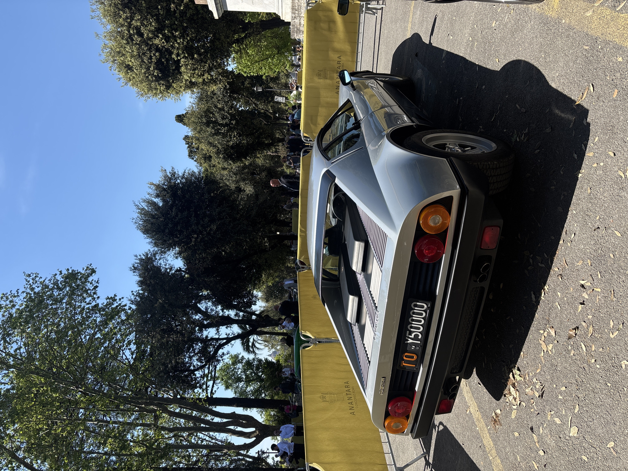 A silver De Tomaso Pantera with the license plate "TO Y50000" is parked on an asphalt surface, casting a shadow to the right. The backdrop features a yellow fence with "ANANTARA CONCORSOROMA" printed on it, as well as trees and a blue sky. People are visible in the background near the fence. The location is implied to be in Rome, Italy, based on the fence's text.