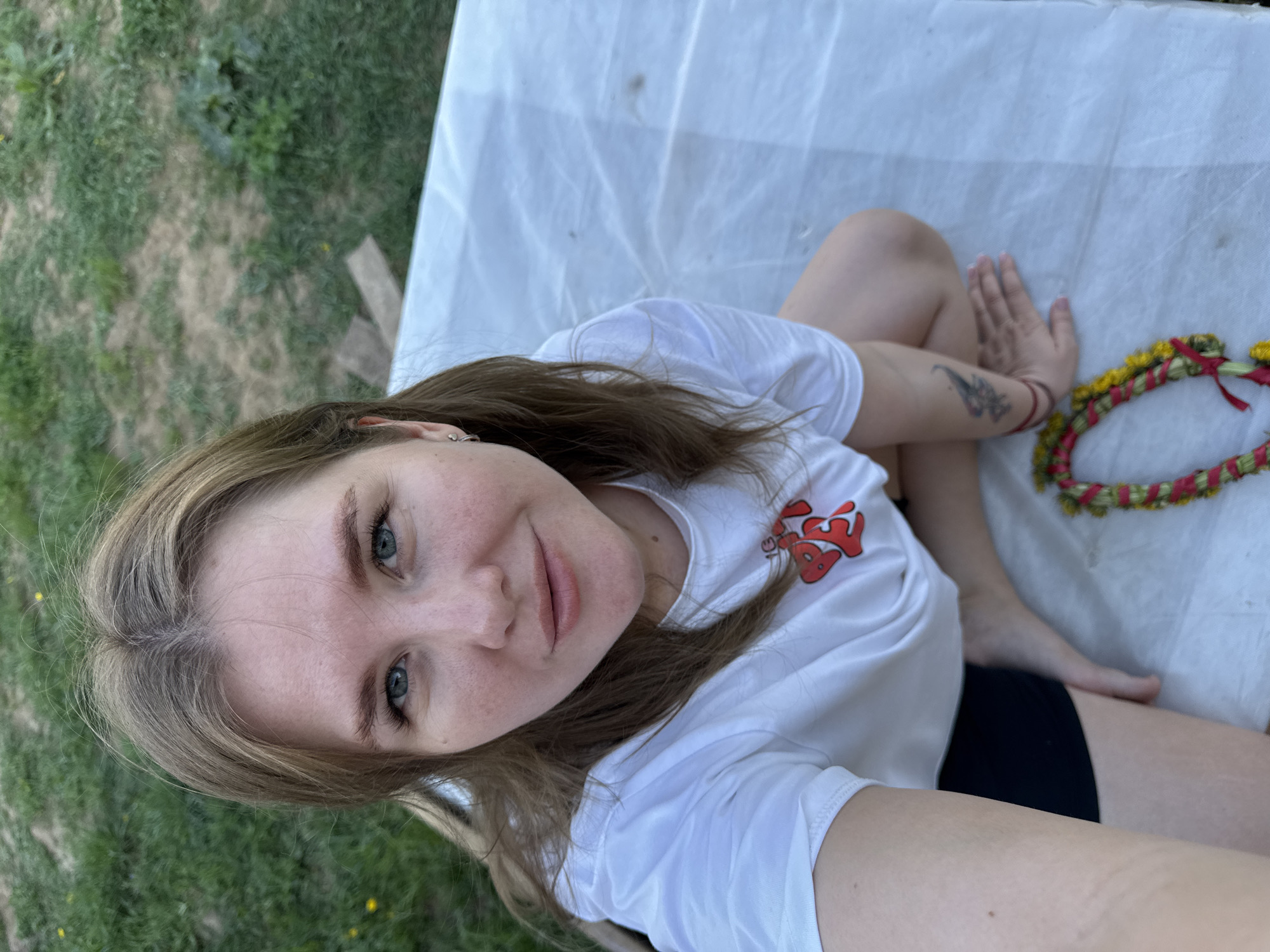 A young woman is sitting cross-legged on a white sheet outdoors. She is wearing a white t-shirt with a red design and black shorts. She has light brown hair and blue eyes. She is looking directly at the camera. A wreath of flowers lies next to her on the sheet. The background is green grass. This image seems to be taken in a garden in Ферма, Russia.