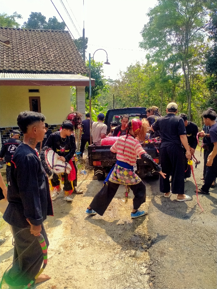 The scene depicts an outdoor gathering on an unpaved road in Tawangsari, Indonesia, under bright daylight. Approximately 15-20 individuals, predominantly adult males, are present, many appearing involved in or preparing for a cultural performance. In the immediate foreground to the left, a barefoot male in dark attire holds a multi-colored coiled rope. Center-right, a performer in a pink and white striped shirt, dark wide-legged pants, and a multi-colored patterned apron is poised in a wide stance, wearing blue shoes and a reddish, dreadlock-like wig. To the left of center, another performer in black holds a white and red hobby horse (Kuda Lumping). A dark-colored pickup truck is parked centrally, its bed loaded with a red drum and other objects, with several individuals gathered around it, seemingly organizing or unloading items. On the left side of the frame, a traditional house with a brown tiled roof and light yellow walls is visible. The background features dense green trees and a street light with a curved arm. Overhead power lines are also visible.