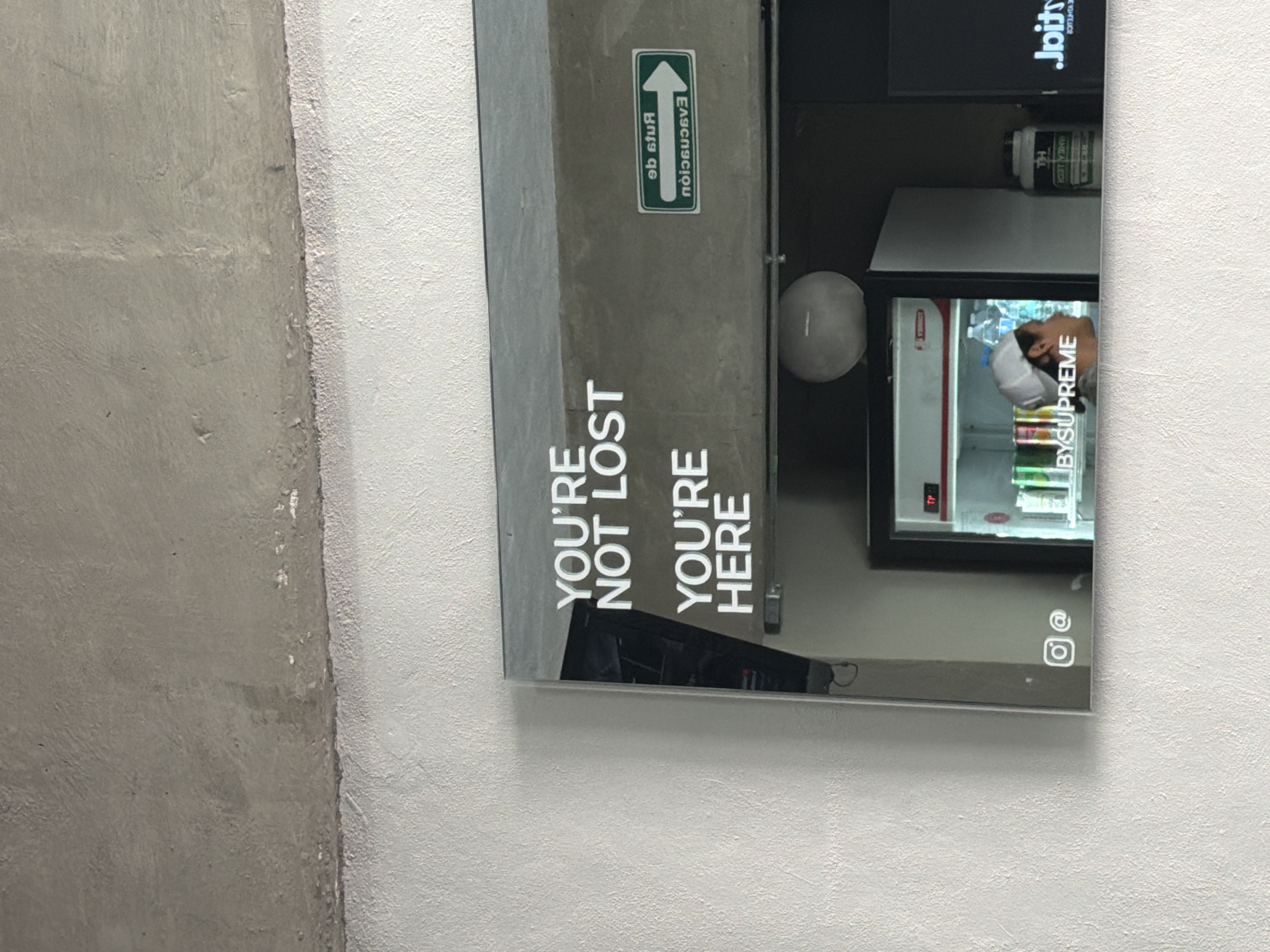 Here's a detailed description of the image:
**Scene Overview**
The image shows a framed mirror mounted on a white wall, reflecting a gym or similar fitness-related environment. The mirror itself has text printed on its surface, and the reflection shows elements of a gym interior.
**Elements in the Mirror/Reflection**
* A small refrigerator with glass door, containing various drinks or snacks.
* A person reflected in the refrigerator door, suggesting someone standing nearby.
* A sign with the text "YOU'RE NOT LOST / YOU'RE HERE".
* A sign with the text "Evacuation route".
* A black face mask.
* An Instagram symbol is printed in the lower right corner of the mirror.
* A container of protein/supplement, and a monitor/display screen are visible in the upper portion of the reflection.
**Wall and Surroundings**
* The mirror is mounted on a textured white wall.
* To the left of the mirror, a rough, gray concrete or plaster wall is visible.
**Location**
Based on the "BYSUPREME" sticker, it is very likely that this photo was taken inside or in front of the gym called Supreme, located in Puebla, Mexico. The photo was taken and the texts in the mirror are mirrored.