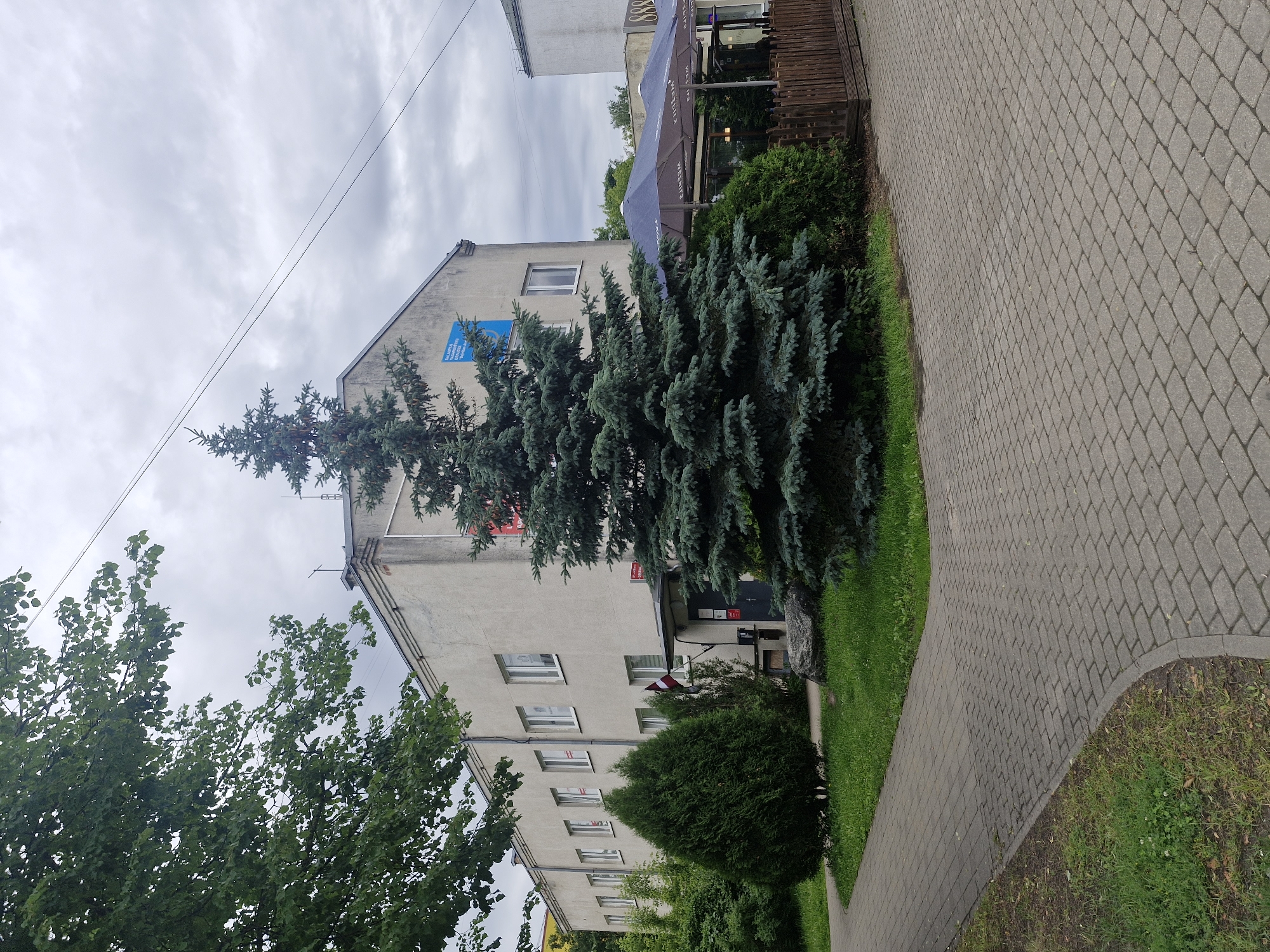 The image shows an exterior view of a multi-story building with a paved walkway in the foreground. The building appears to be made of concrete and has several windows. There is greenery in front of the building, including trees and bushes. A blue sign with white text is visible on the side of the building, as is a Latvian flag. An umbrella and some outdoor seating are also visible near the building. The sky is cloudy, suggesting overcast weather. The paved walkway is made of interlocking stones and curves around a patch of grass. The overall scene appears to be a public space, possibly in a town or city.