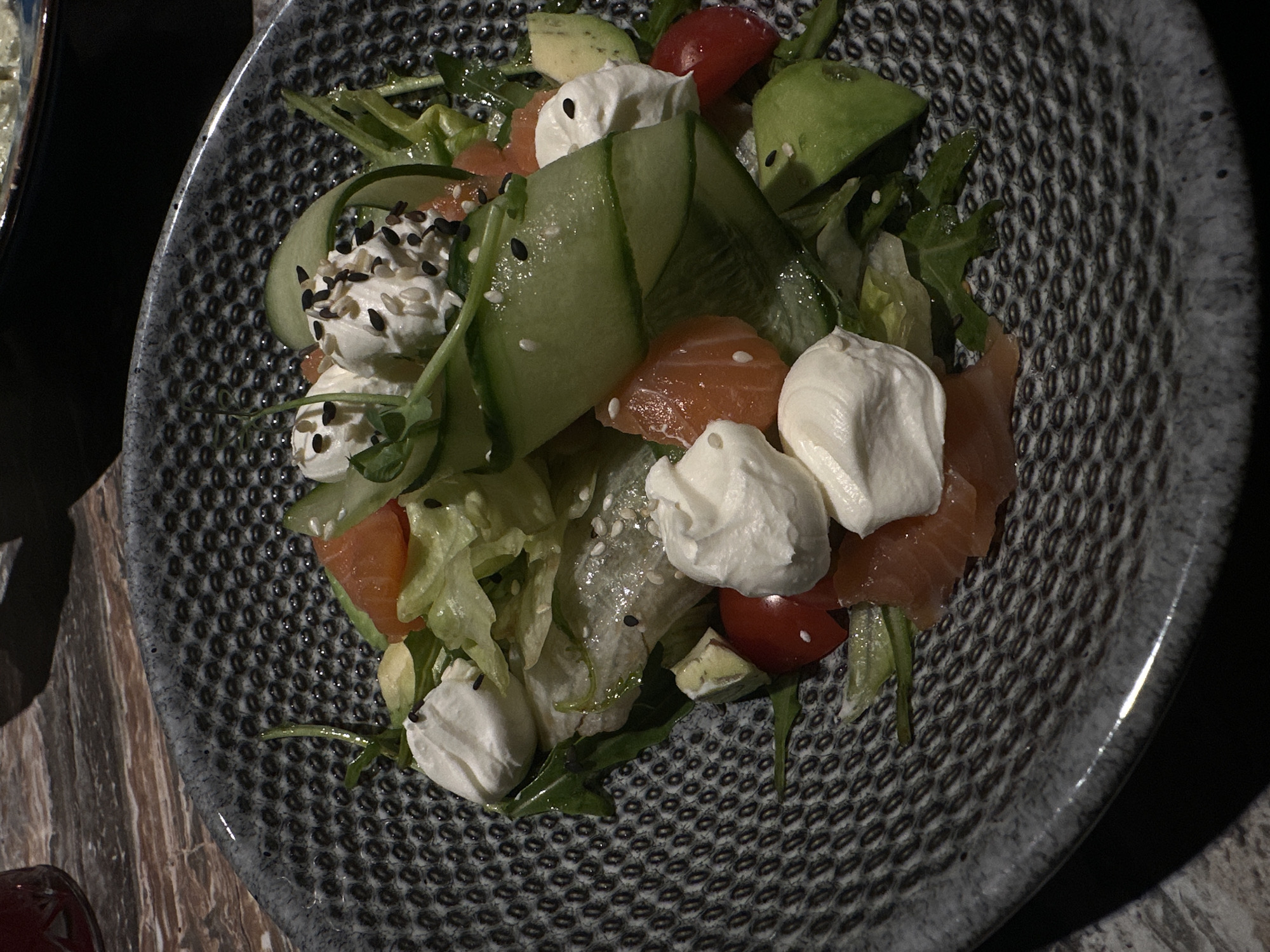 The image shows a close-up view of a salad served on a patterned grey plate. The salad contains ingredients such as salmon, avocado, cucumber ribbons with sesame seeds, small tomatoes, lettuce, and dollops of white cream cheese also topped with sesame seeds. The plate sits on a table, with a glimpse of another dish visible on the left edge of the frame and a red object at the bottom left. The lighting creates shadows, giving the scene a slightly moody atmosphere. There is no indication of the image's location within the city of Chaikovsky, Russia, or any further details about people or events.