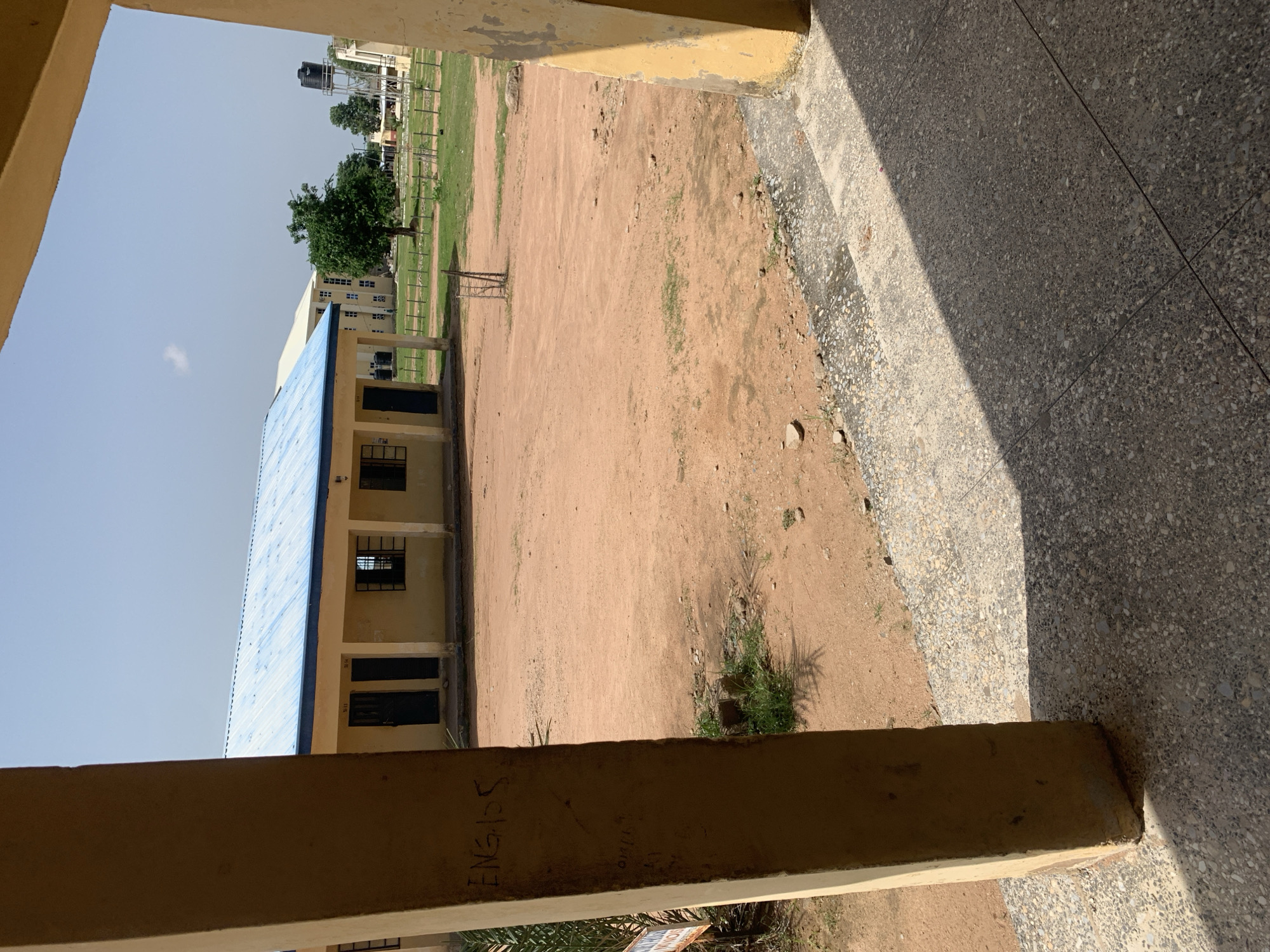The image depicts an outdoor scene in Jalingo, Nigeria, likely a school or educational institution. In the foreground, a pale yellow pillar of a building is visible on the left, with faint graffiti, including the word "ENGISS," written on its surface. To the right, a paved walkway with a speckled grey finish is seen, casting a long shadow from the pillar.
Beyond the immediate foreground, a dirt courtyard stretches out, sparsely covered with patches of green grass. A single-story building with a blue corrugated metal roof and yellow walls occupies the middle ground. This building has several dark blue doors and windows, some with bars. The numbers "30-13" and "30-14" are visible above two of the doors, suggesting it might be a block of classrooms or offices.
Further in the background, other buildings with yellow walls and white roofs are visible, along with mature trees. A water tower on stilts stands to the right of the buildings. The sky is a clear, light blue with a few faint clouds. The overall impression is of a functional, perhaps slightly aged, educational complex under a bright, sunny sky.