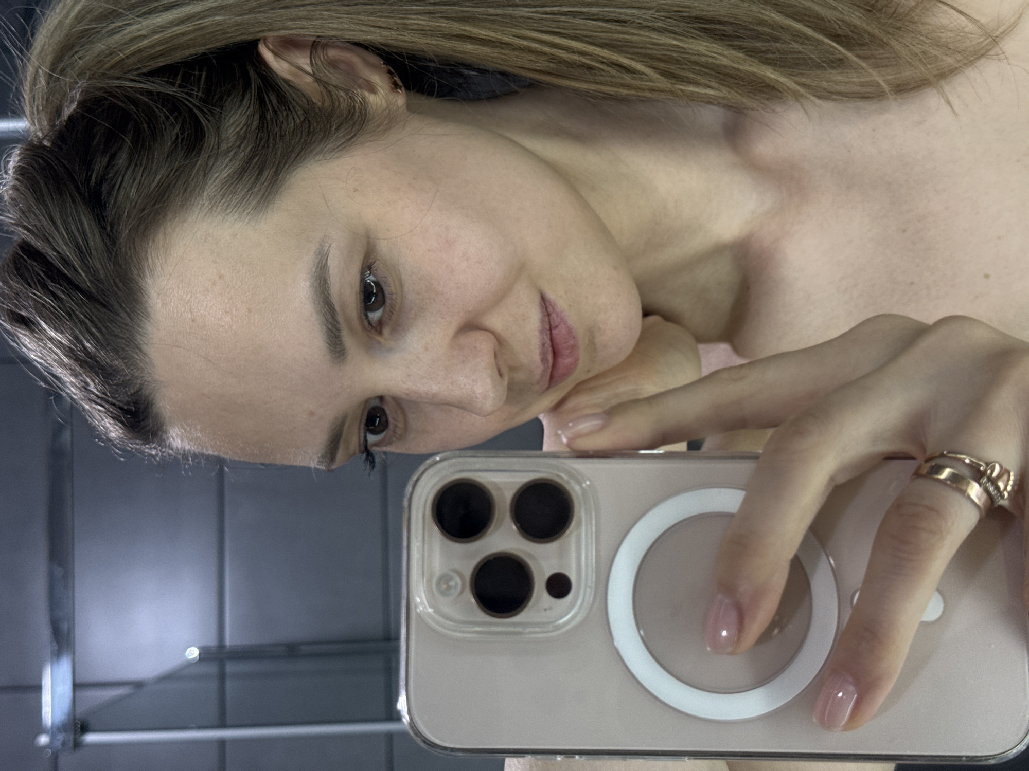 A fair-skinned woman with brown hair is taking a selfie with her iPhone. The phone has a clear case and MagSafe ring. She is wearing two gold rings on her right ring finger. The background is a gray tile wall. There is no evidence to suggest the location is in Chaikovsky, Russia.