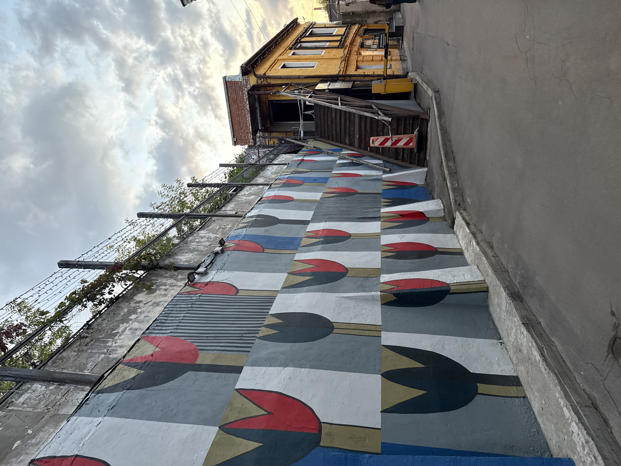 The image depicts a narrow alleyway in Moscow, Russia, alongside a building adorned with a colorful mural of stylized tulips. The mural, featuring bold red and black tulip shapes against a background of grey, white, and blue geometric blocks, covers a sloped concrete wall. Above the wall, barbed wire and leafy branches are visible against a cloudy sky.
To the right of the mural, a two-story yellow building stands, featuring an exterior staircase leading to the second floor. The building has several windows and appears to be an establishment with a sign that reads "ВНИМАНИЕ!" (Attention!) in Russian. A yellow barrier gate is positioned near the entrance of the building.
The alleyway itself is paved with asphalt, showing some cracks. A red and white striped traffic cone is placed at the base of the stairs. A few people are partially visible in the scene, one on the right side of the alley and another near the yellow building's entrance. The overall impression is one of an urban, artistic space with a somewhat industrial feel.