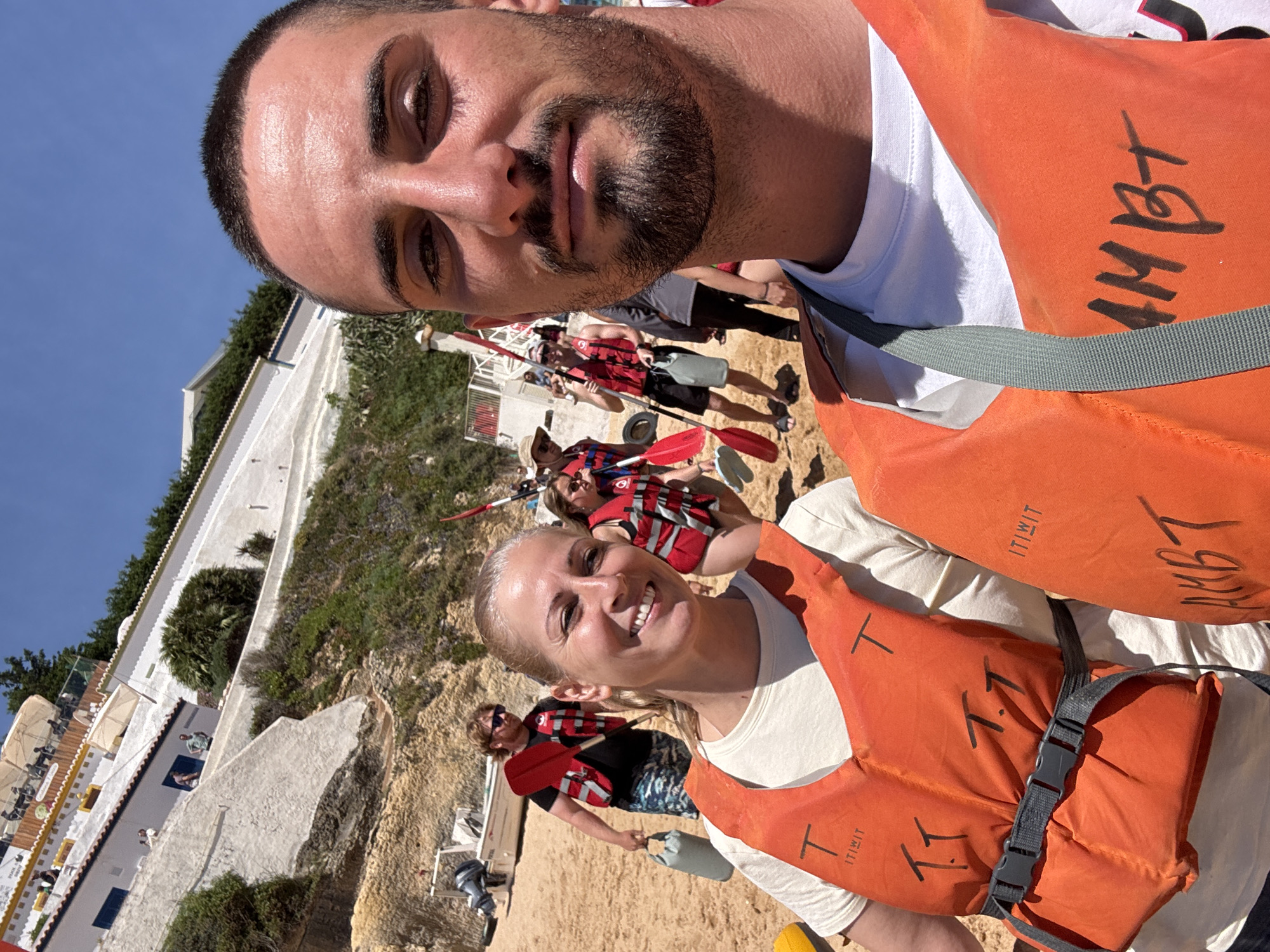 A selfie shows a man and a woman on a beach in Unknown, Unknown. Both are wearing orange life vests over white t-shirts. Behind them, a group of people is also wearing life vests and holding paddles. The background features a white building, vegetation, a sandy beach, and a clear blue sky.