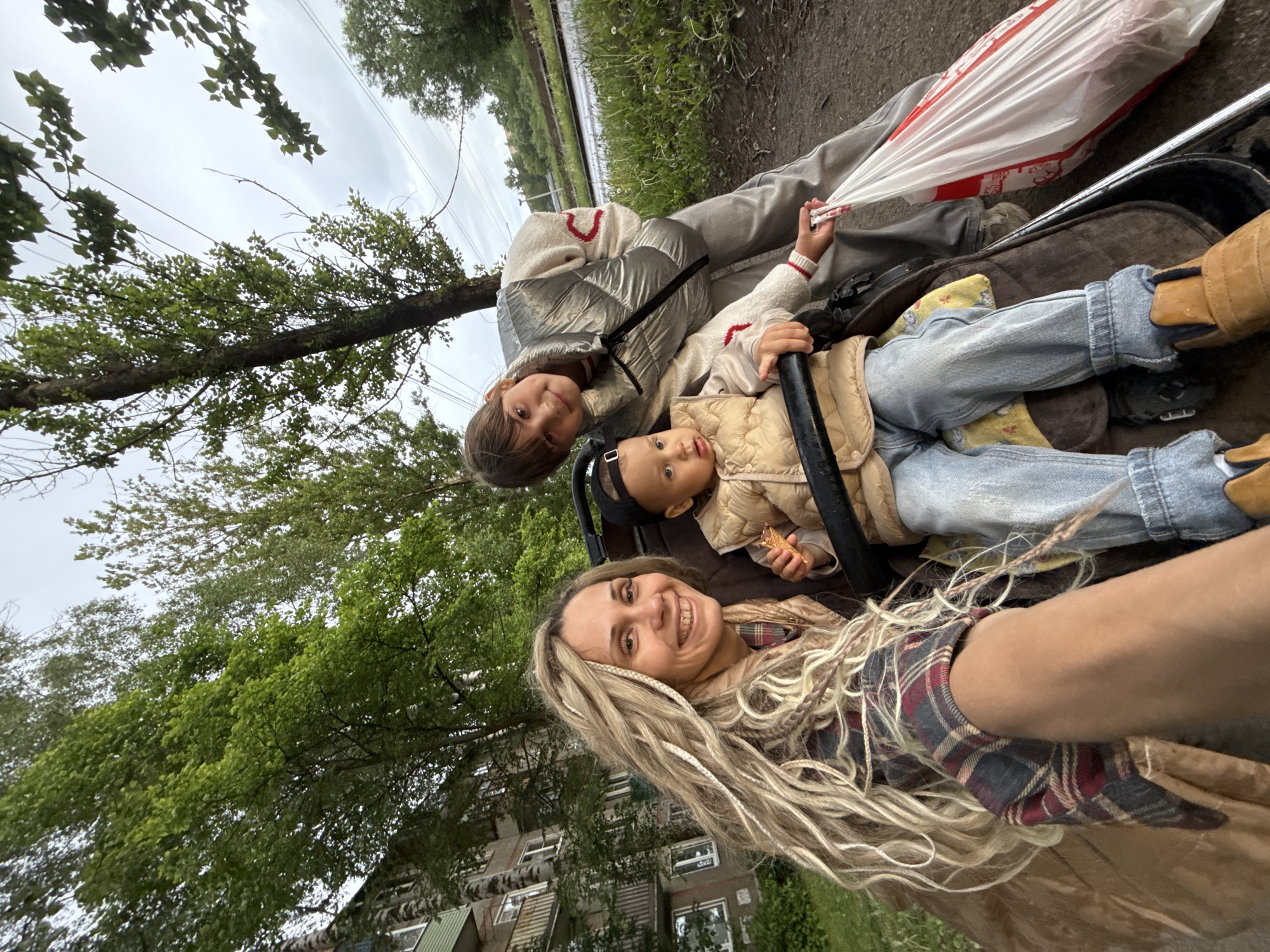 The image depicts a woman with blonde dreadlocks taking a selfie with two children in a stroller. The stroller is positioned on a pathway next to a grassy area and a building with multiple windows. The three are outside in what appears to be the city of Rusanovo, Russia. The sky is overcast, suggesting it is a cloudy day. The elder child is positioned to the left of the younger child, both are in the stroller. The adult is holding up their right arm to take the photo. The younger child is holding food in their left hand.