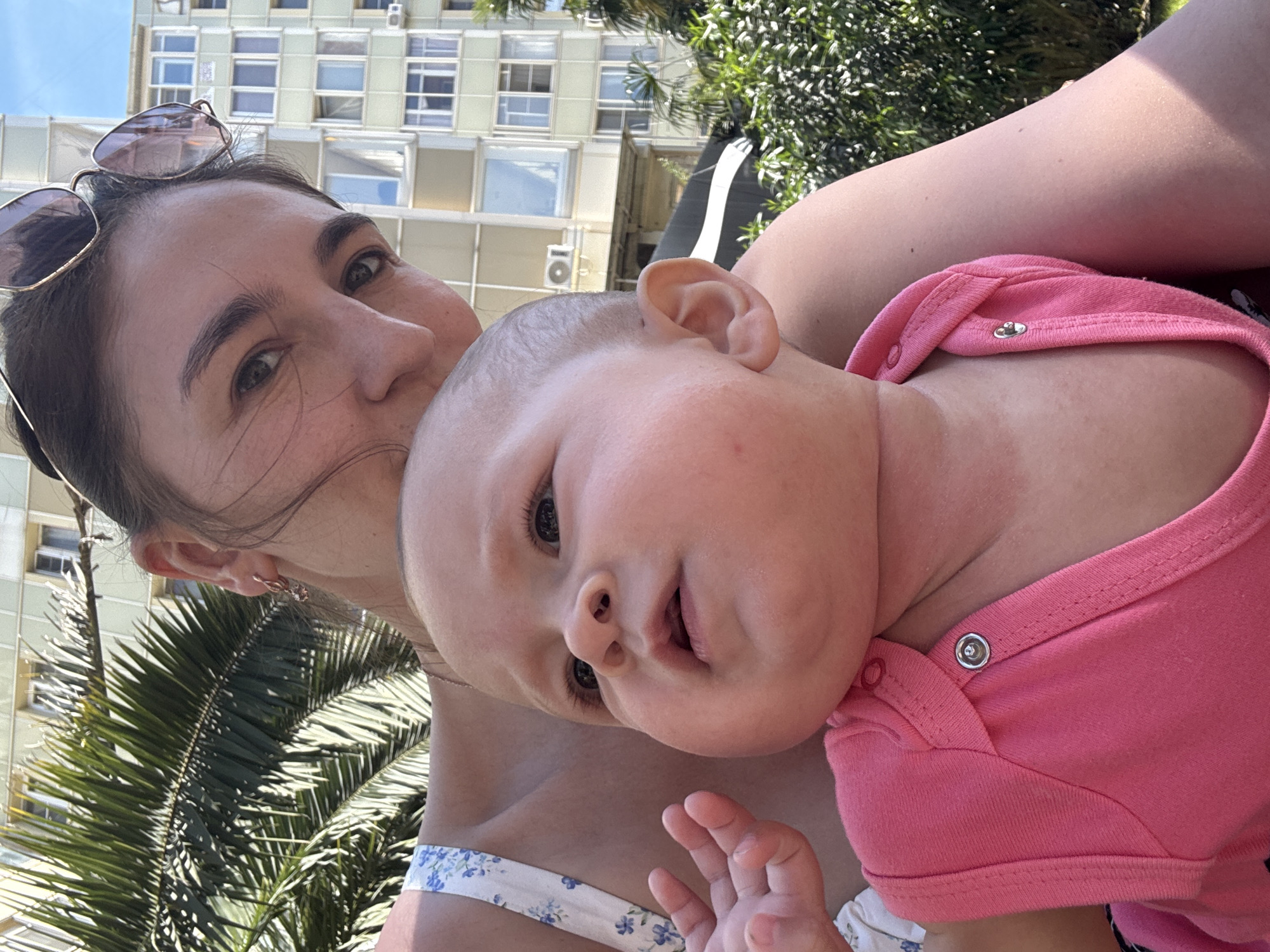 A close-up, selfie-style photograph depicts a woman holding a baby outdoors on a bright, sunny day. The woman, positioned on the left, has dark hair pulled back with sunglasses resting on her head. She wears a light-colored top with a blue floral pattern and looks towards the baby or the camera with a gentle expression. The baby, held close to the woman, has fair skin, light hair, and is dressed in a vibrant pink onesie. The baby's gaze is directed towards the viewer with a curious, slightly open-mouthed expression.
The background features a multi-story, light-colored building with numerous windows, suggesting an urban or resort setting. Lush green foliage, including prominent palm fronds, is visible around the building and in the foreground, indicating a warm, possibly subtropical climate. The strong sunlight and clear skies confirm it is daytime, likely midday or early afternoon. The overall mood of the image is warm, tender, and happy, capturing a loving moment between a mother and her child. This scene is consistent with the characteristics of Sochi, Russia. No visible text is present.
