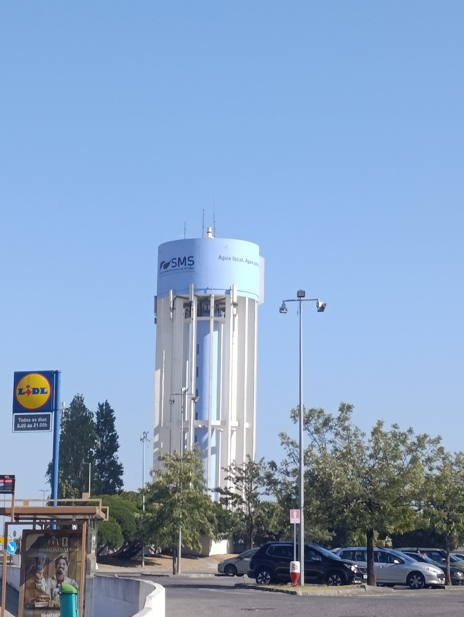This is a medium-shot, eye-level outdoor photograph taken on a clear, sunny day, likely in the afternoon given the bright lighting. The central subject is a tall, cylindrical water tower, predominantly white with a blue band at the top. The blue band features the logo "SMS" and the Portuguese text "Água local. Água pública." (Local water. Public water.). To the left of the water tower, a yellow and red Lidl supermarket sign stands, indicating the store's opening hours: "Todos os dias 8:00 às 21:00h" (Every day 8:00 AM to 9:00 PM). In the foreground, there's a billboard advertising a "Mundial" product with a man holding a burger. A blue recycling bin is visible next to it. A street lamp with multiple lights stands to the right of the water tower. Several cars are parked in a lot, with trees and foliage scattered throughout the scene. The overall setting appears to be a commercial area or a roadside location in Setúbal, Portugal. The clear blue sky and bright sunlight suggest pleasant weather. There are no discernible emotions conveyed by any subjects in the image.