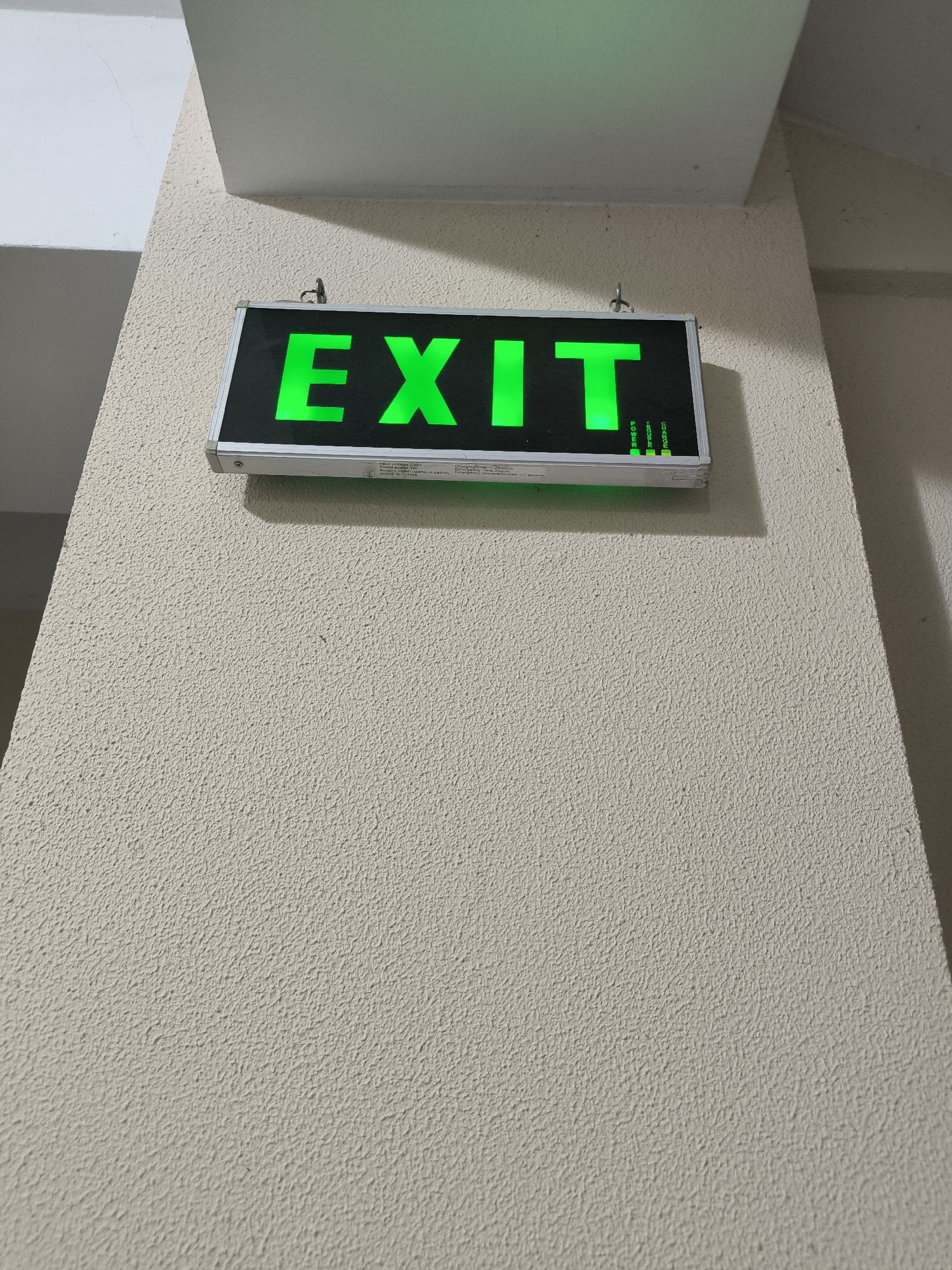 An illuminated green "EXIT" sign is mounted on a textured, light-colored wall. The sign is rectangular with a silver metal frame and the letters "EXIT" are bright green and luminescent. Below the letters, there are smaller labels and indicator lights. The wall has a rough, stucco-like texture. To the left of the sign, a portion of a white, angled ceiling or architectural feature is visible. The environment appears to be indoors, possibly in a public building or hallway. No people are present in the image. The specific location in Chișinău, Moldova cannot be determined from the visual information provided.