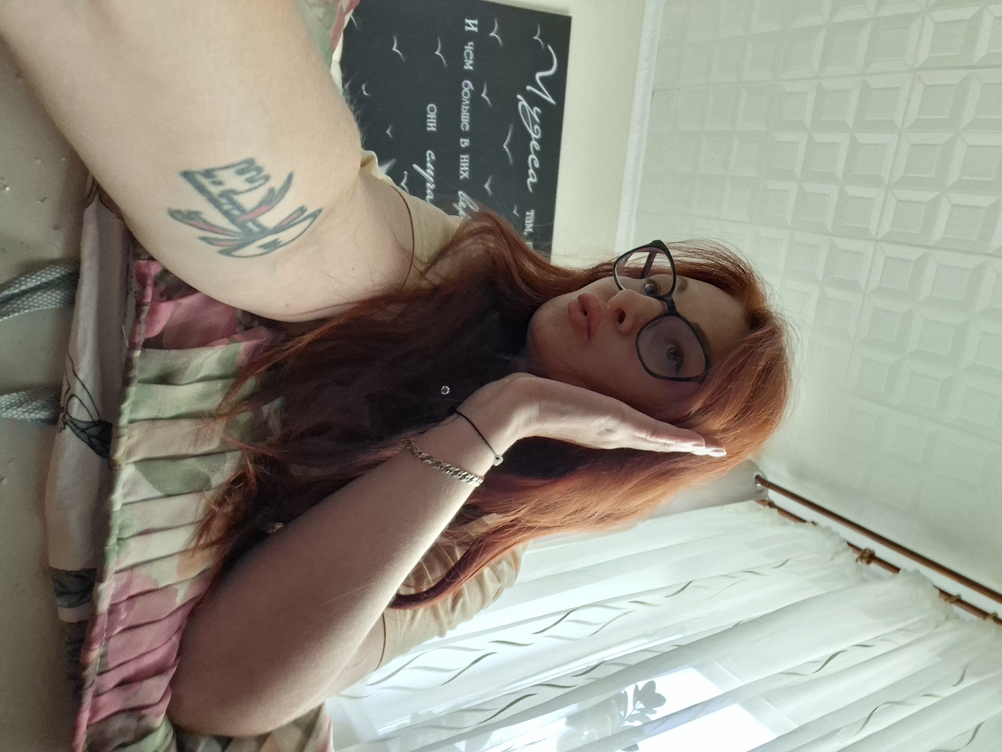 A young woman with long, wavy red hair and black-rimmed glasses is shown indoors, in what appears to be a bedroom in Myadzyel, Belarus. She is lying down on a bed with a patterned duvet, with her left arm resting on the bed and her right arm raised to her face, her hand touching her temple. She is wearing a tan t-shirt. A tattoo is visible on her left forearm, and a thin bracelet is on her right wrist. Behind her, a dark chalkboard with white lettering and bird silhouettes is mounted on the wall. The text on the chalkboard appears to be in Russian. A white sheer curtain with a floral pattern is visible in the background, near a window. The ceiling is white with a textured, geometric pattern.
