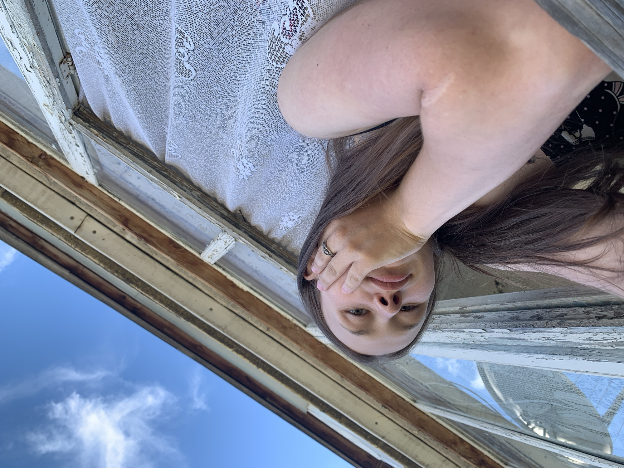 The image shows a young woman with long brown hair looking up at the camera from an unusual low angle. She is resting her cheek on her hand and wearing a ring on her finger. She is positioned outdoors next to a weathered wooden window frame with peeling paint. A white lace curtain is visible behind her. The sky above is blue with wispy white clouds. The scene appears to be located near a building with a rustic, aged appearance. While the image does not explicitly state the location within Фоки, Russia, the overall aesthetic suggests a rural or older urban setting typical of many locations in Russia.
