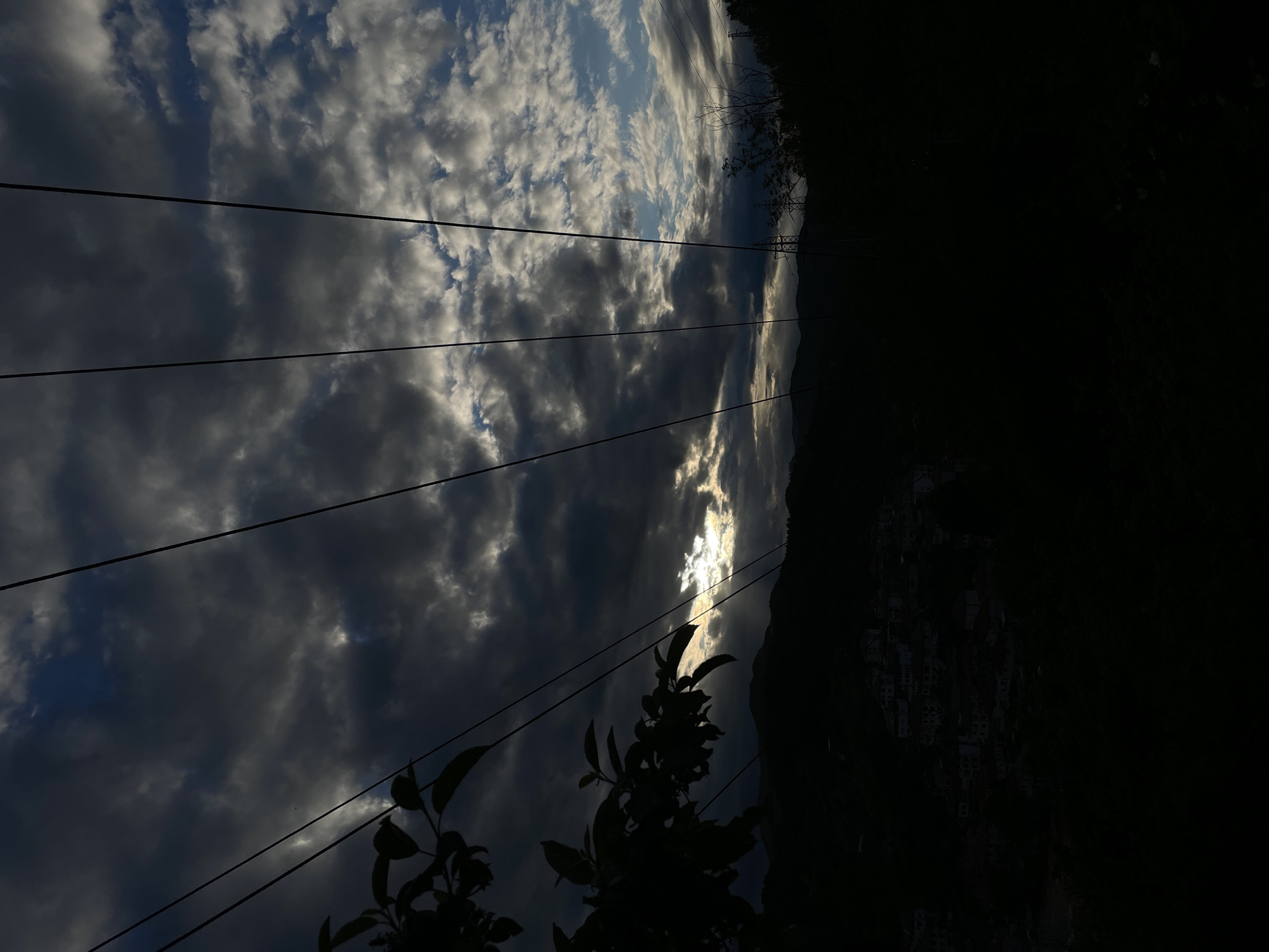 The image shows a view of the sky with scattered clouds and power lines in the foreground. The sky has varying shades of gray and some patches of blue. The sun is partially visible through the clouds, creating a bright glare. On the right side of the image, a dark mass suggests a hillside or mountain, with some vague indication of structures or buildings. The scene is likely in Zlatograd, Bulgaria, given the location provided. Vegetation, possibly trees or bushes, is silhouetted in the lower part of the image.