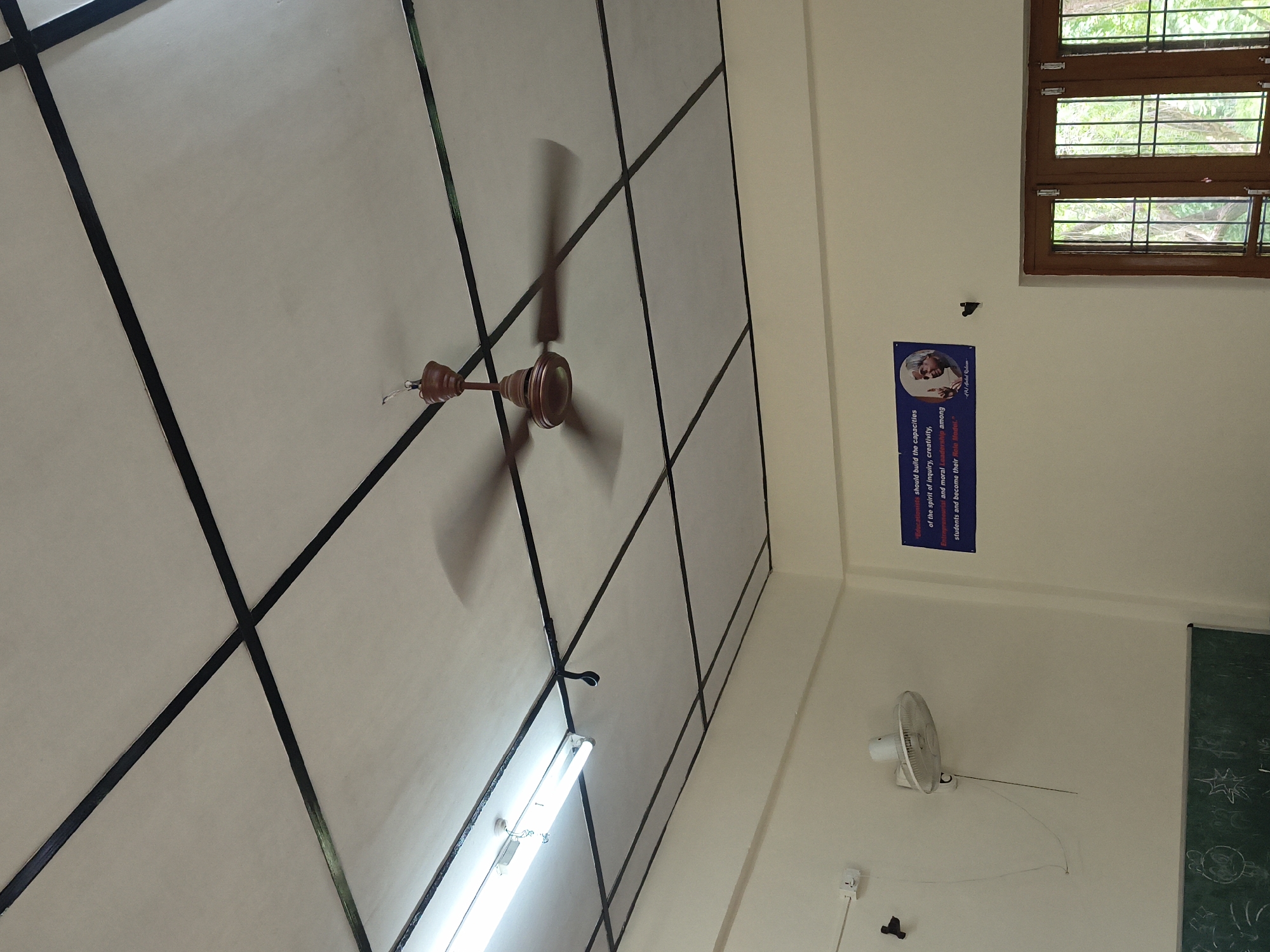 The image shows the interior of a room, likely a classroom or lecture hall. A ceiling fan is in motion, its blades blurred. The ceiling is white with a grid pattern of black lines. A fluorescent light fixture is also visible on the ceiling. On the wall, a white wall-mounted fan is present, and below it, a portion of a chalkboard with drawings is visible. To the right, a blue poster with a quote and an image of a man is displayed on the wall. A wooden door with glass panes is also visible on the right side of the frame, showing greenery outside. The walls are painted a light cream color.