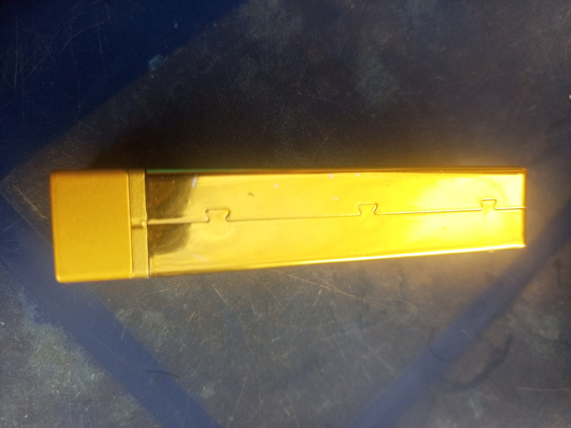A rectangular, metallic gold cigarette case is positioned horizontally on a dark, scratched surface, casting a shadow. The cigarette case has a visible hinge mechanism. The image appears to be taken indoors. There is no discernible connection to Lhokseumawe, Indonesia, in the visual information provided.