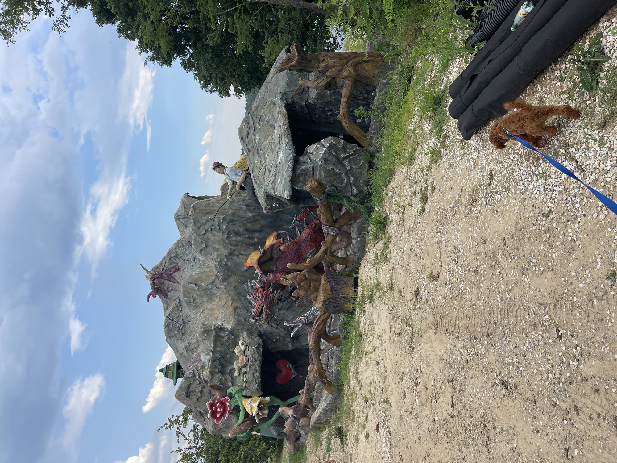 This image captures an outdoor scene in Koprinka, Bulgaria, featuring an elaborate artificial rock formation adorned with fantasy-themed sculptures, a dog, and various environmental elements under a partly cloudy sky.
The central feature is a large, multi-level artificial rock structure, painted in shades of grey and brown to resemble stone and earth. On the left side, a large red and yellow sculpted flower with green foliage emerges from the base of the rock. Beneath an overhang in this section, a prominent red heart sculpture is visible within a dark recess. To the right of the heart, a large red dragon sculpture with golden accents on its wings and head emerges from a cave opening, its head turned towards the left. Below the dragon's head, another creature, possibly a fish or serpent, with an open mouth showing pointed teeth, is sculpted. Integrated into the base of the rock formation are several sculpted tree roots and truncated stumps.
Above the main structure, on the central peak, a figure wearing a green pointed hat is partially visible. Further to the right, on a lower section of the rock, a seated male figure in a white shirt and red cap is depicted holding a fishing rod, facing towards the right. Another abstract, possibly bird-like, red sculpture sits atop the highest peak on the right.
The foreground consists of a dirt and gravel path with visible tire tracks. On the far right of the foreground, a small, reddish-brown curly-haired dog on a blue leash walks towards a stack of black corrugated pipes. A green plastic bottle is visible on the ground next to the pipes.
In the background, on the left, dense green foliage is visible. On the right, numerous green trees extend upwards, partially obscuring a body of water which is faintly visible beyond them. The sky is light blue with scattered white cumulus clouds.