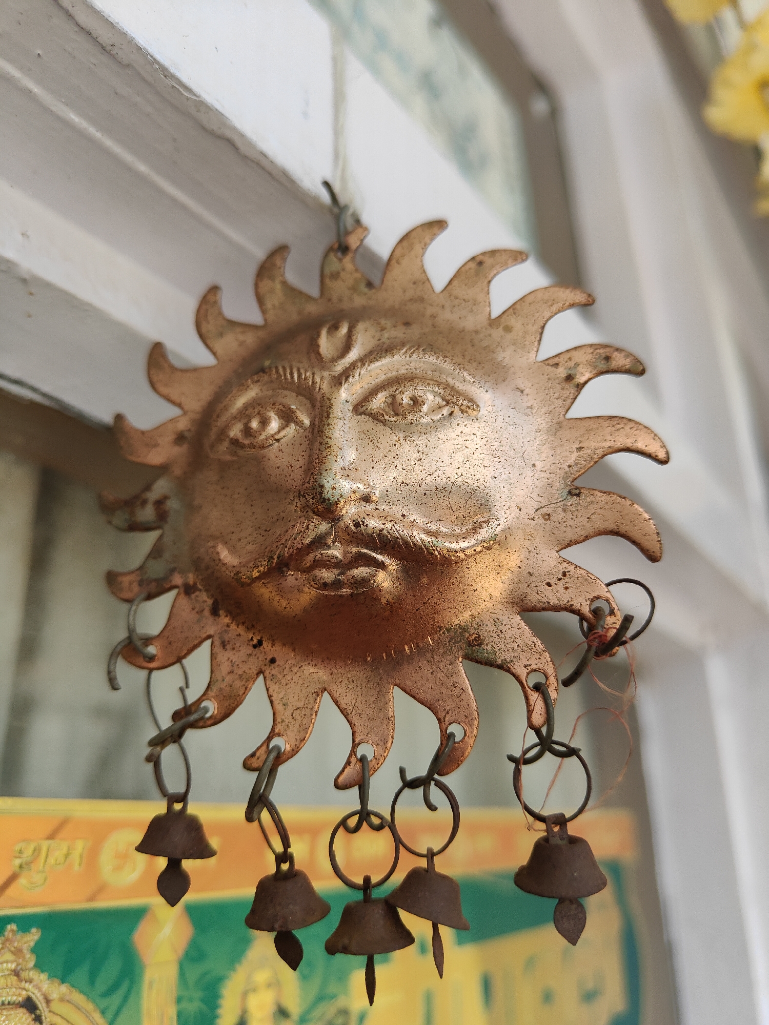 A weathered, copper-toned sun-shaped wind chime hangs from a white wooden beam, likely in Vashisht, India. The sun face is detailed with eyes, a nose, a prominent mustache, and a third eye marking on its forehead. Rays emanate from the sun's disc, each with a small hole from which small, bell-shaped chimes hang via metal rings. The chimes are a darker, oxidized metal and have a simple, conical shape with a small leaf-like adornment at the bottom. The background shows hints of a colorful poster or artwork with green and yellow hues, possibly depicting religious imagery, and a blurred glimpse of a yellow object, potentially flowers, in the upper right corner. The overall impression is of a decorative item exposed to the elements, with some signs of age and patina.