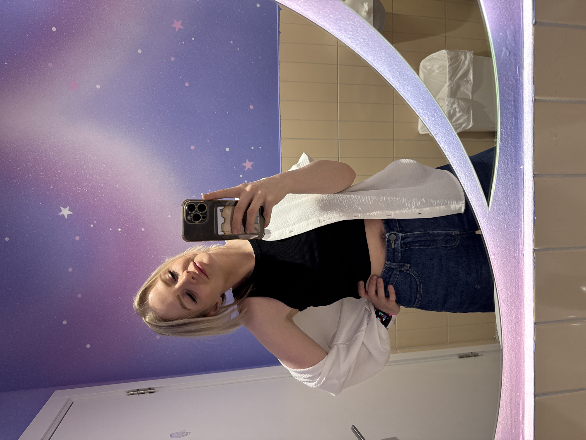 A young blonde woman is depicted taking a mirror selfie in an indoor setting, likely in Saint Petersburg, Russia. She has fair skin and straight blonde hair, framing her face. She is dressed in a black fitted top and blue denim jeans, with a white, crinkled-texture long-sleeved shirt or jacket draped over her shoulders. A smartphone in a dark case is held in her right hand to capture the reflection, while her left hand rests casually near her hip, revealing a smartwatch with a colorful display on her wrist.
The background behind her in the mirror features a distinctive wall adorned with a purple and pink gradient, embellished with white and pink star decals and speckles, creating a celestial or galaxy theme. To the right of the reflection, a section of light beige tiled wall is visible, on which a white, plastic-covered object, possibly a trash receptacle, is mounted. A white door is also reflected on the far left. The mirror itself appears to have an illuminated frame, casting a soft, diffused light. The woman looks towards her phone screen with a slight, closed-lip smile, conveying a relaxed mood. No prominent text is clearly visible, though a faint "Mon" can be seen on the phone's reflection. The scene suggests a modern and whimsically decorated public or commercial space.