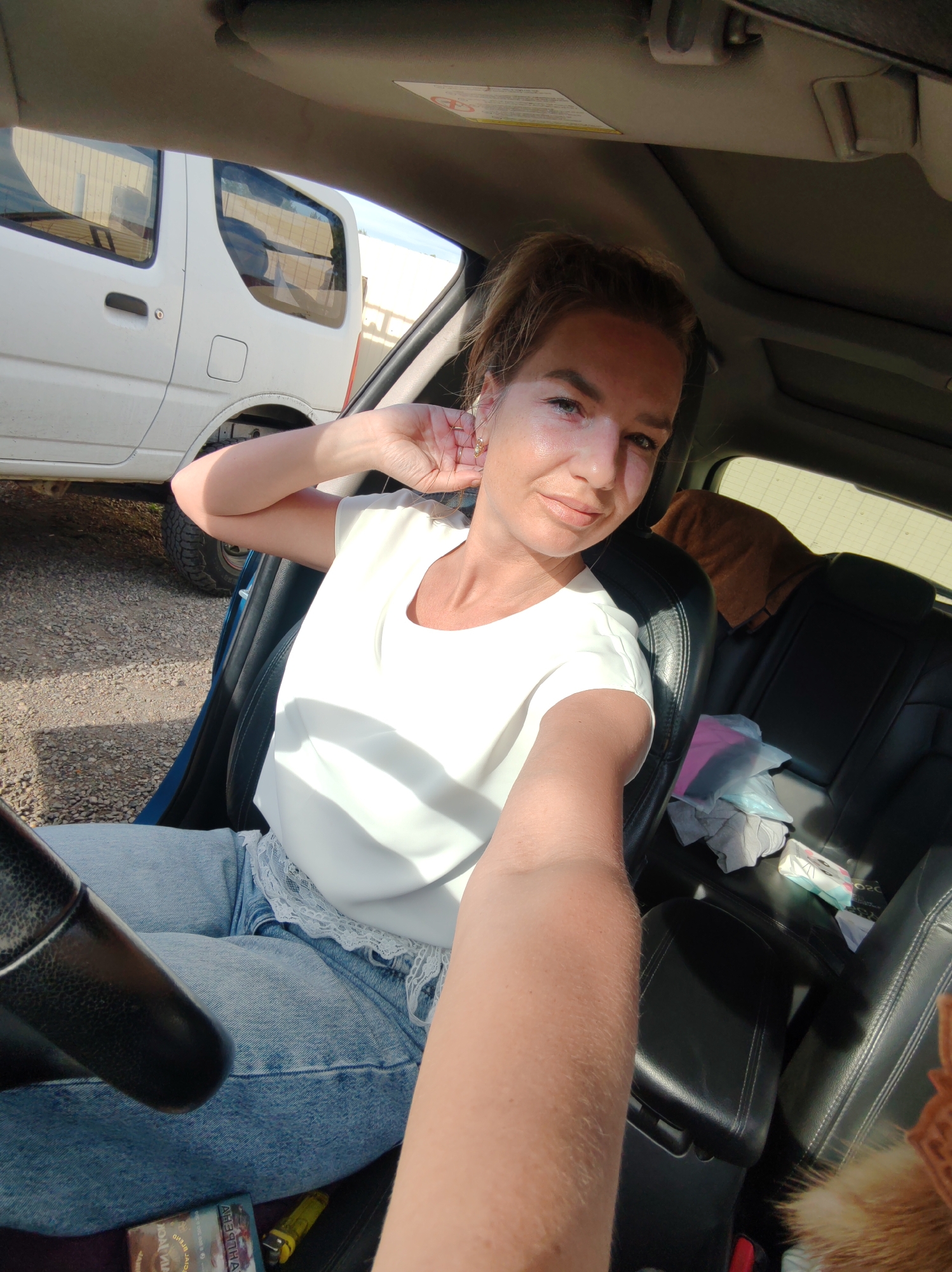 The image shows a woman sitting in the driver's seat of a car. She is wearing a white top and blue jeans. Her left arm is resting on the steering wheel. The car appears to be parked on a gravel lot. In the background, another white vehicle is visible. The scene is outdoors, with sunlight casting shadows. The image was taken in Paratunka, Russia.