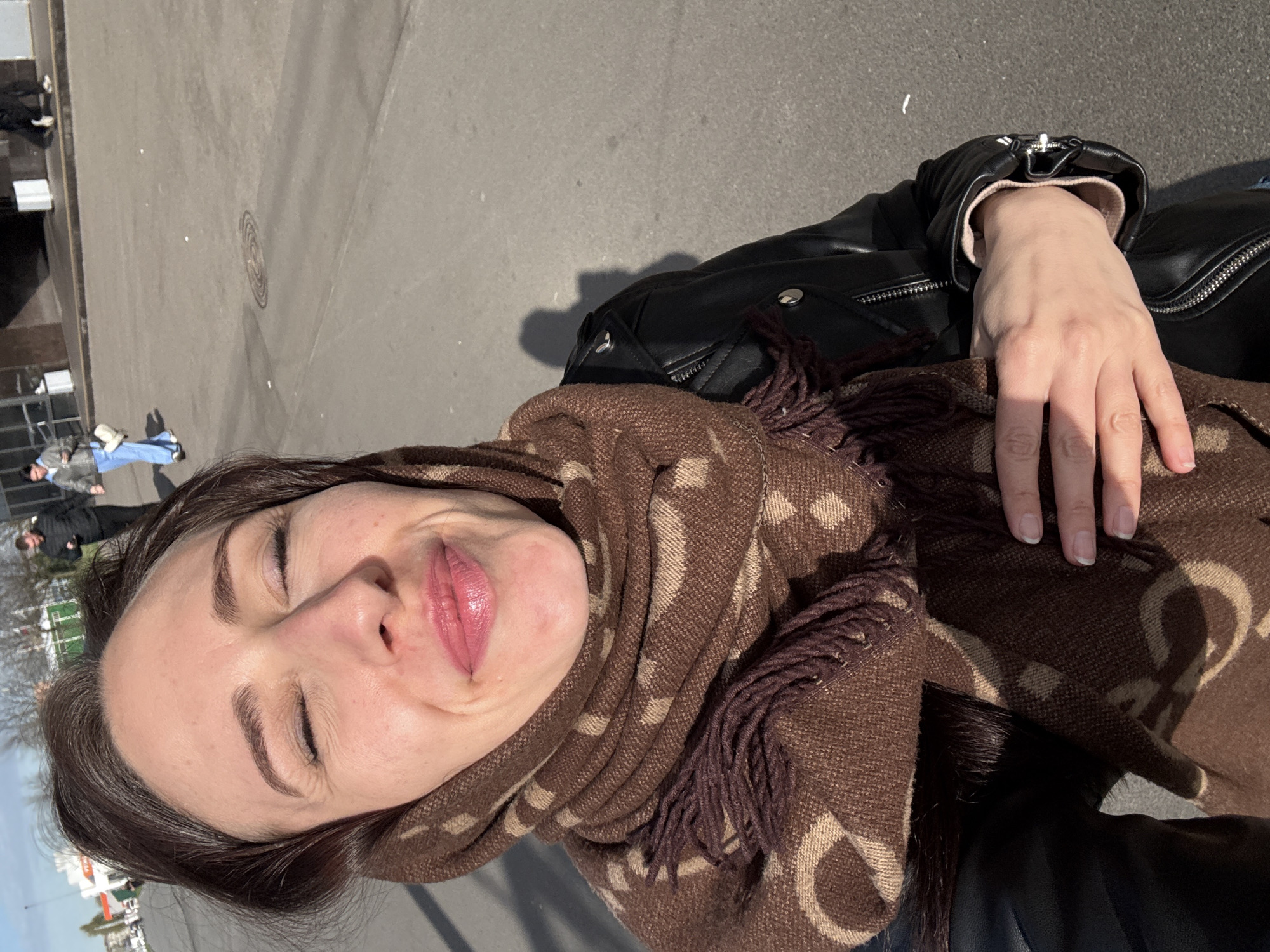 A woman with dark hair and closed eyes is seen in an outdoor shot, possibly a selfie. She is wearing a brown patterned scarf and a black leather jacket. Her hand rests on the scarf. The background appears to be a city street with people and buildings. Based on the architectural details and street layout visible in the background, it is likely that the location is Saint Petersburg, Russia.