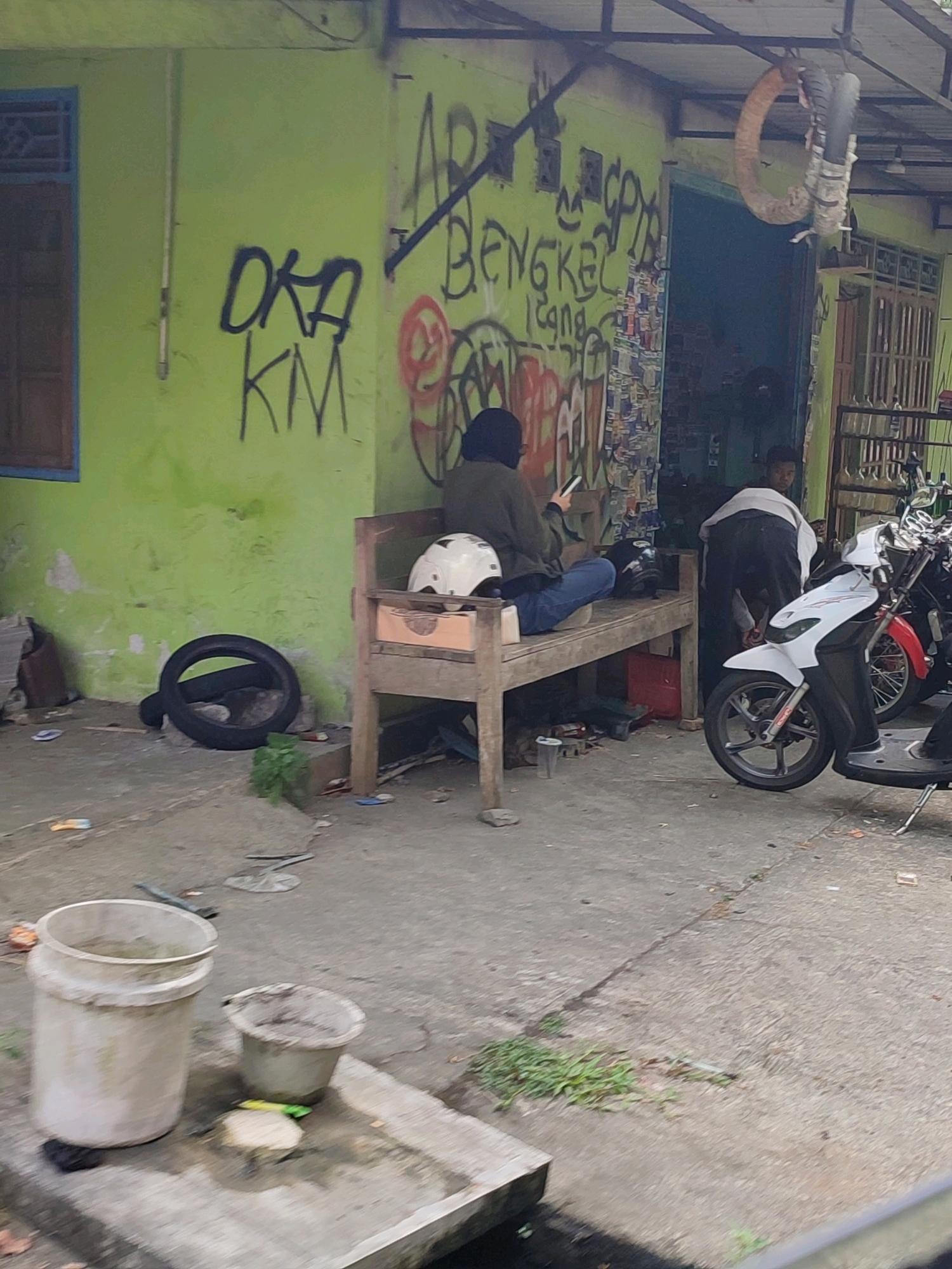 The image depicts an outdoor scene in front of a building with a bright green wall covered in graffiti. A person wearing a dark hijab and a green jacket is seated on a wooden bench, looking at a mobile phone. Beside them on the bench is a white helmet. In front of the bench, a black tire is leaning against the wall. To the right of the bench, a white and red motorcycle is parked. Another person is partially visible behind the motorcycle, appearing to be tending to something within the building's entrance. In the foreground, two large buckets, one white and one a lighter shade, are visible on the concrete ground. The environment appears somewhat cluttered with debris on the ground. The graffiti on the wall includes text like "OKA KM" and other illegible scribbles. The scene suggests a roadside workshop or a similar establishment.