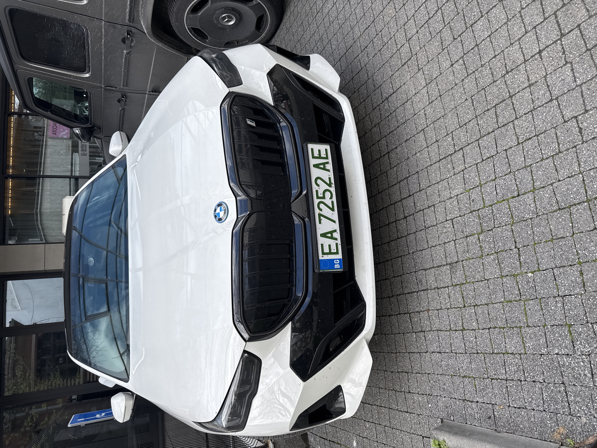 The image shows a white BMW car with a Bulgarian license plate "EA 7252 AE" parked on a cobblestone surface. A gray Mercedes-Benz G-Class is partially visible next to it. The building behind the cars has a glass facade. The license plate indicates the car is registered in Sofia, Bulgaria. Without additional context, determining the exact location within Plovdiv is impossible, but the scene suggests a modern urban environment.