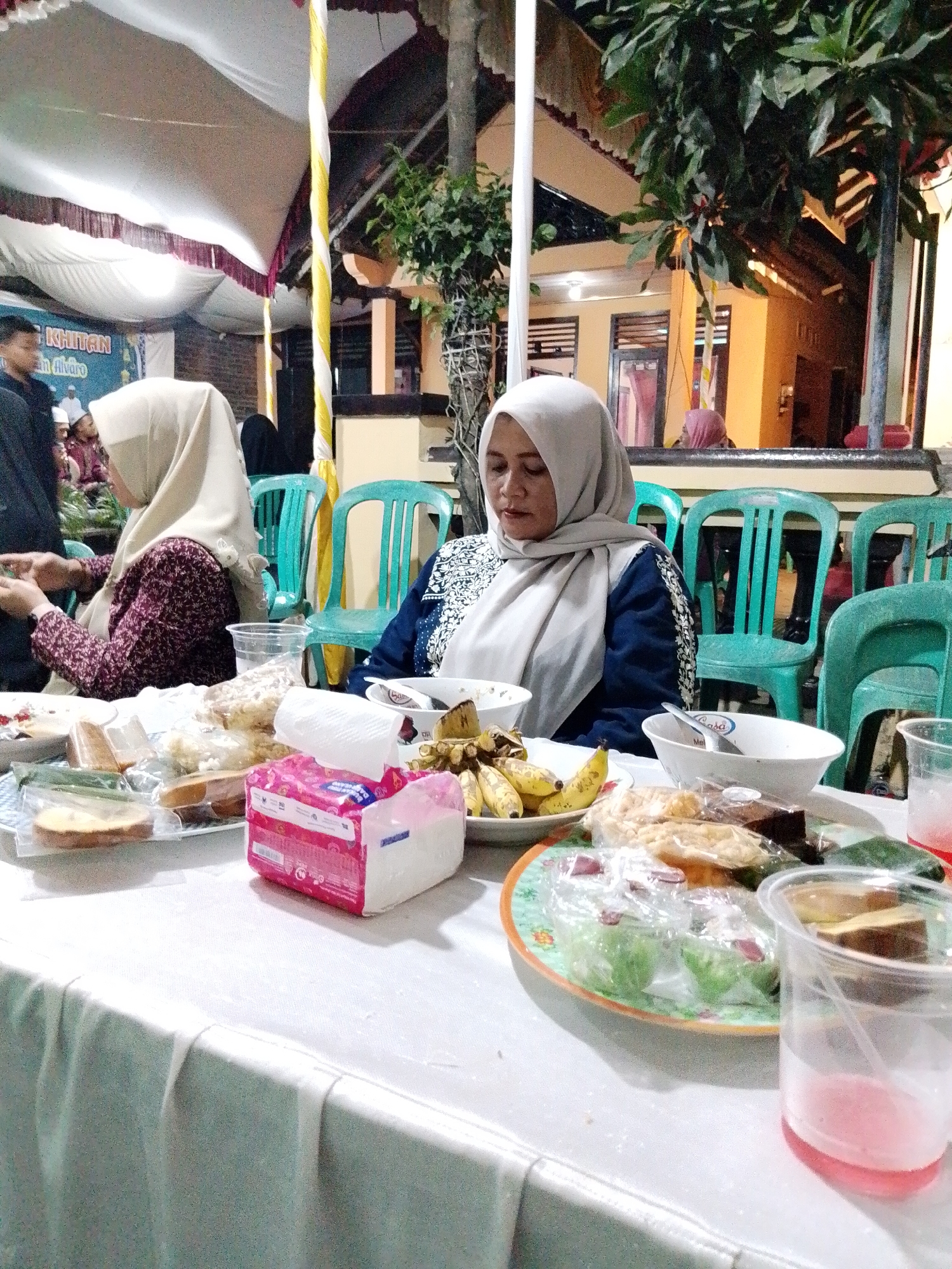 The image depicts an outdoor event, likely a gathering or reception, set up under a white tent with maroon trim. A banner with text in an Indonesian script is partially visible on the left. The foreground is dominated by a white tablecloth laden with food and drinks. Visible items include a stack of white napkins in a pink box, several plates of food containing what appear to be pastries, snacks, and possibly some savory items wrapped in plastic. Bananas are also present on a plate. There are several plastic cups, one containing a pink liquid and a straw.
Two women are seated at the table. The woman in the foreground, on the right, wears a light-colored hijab and a blue and white patterned top. She is looking down at the table. To her left, another woman, also wearing a hijab and a patterned maroon and white top, is actively engaged with her phone. In the background, a row of green plastic chairs is visible, suggesting more seating for guests. Behind the chairs, part of a building with yellow walls and dark-framed windows is seen, indicating the location is likely a residential or community area. The overall atmosphere suggests a communal event, possibly a religious celebration or a family gathering. The presence of the banner and the general arrangement of the scene point to an event taking place in Trangkil, Indonesia.