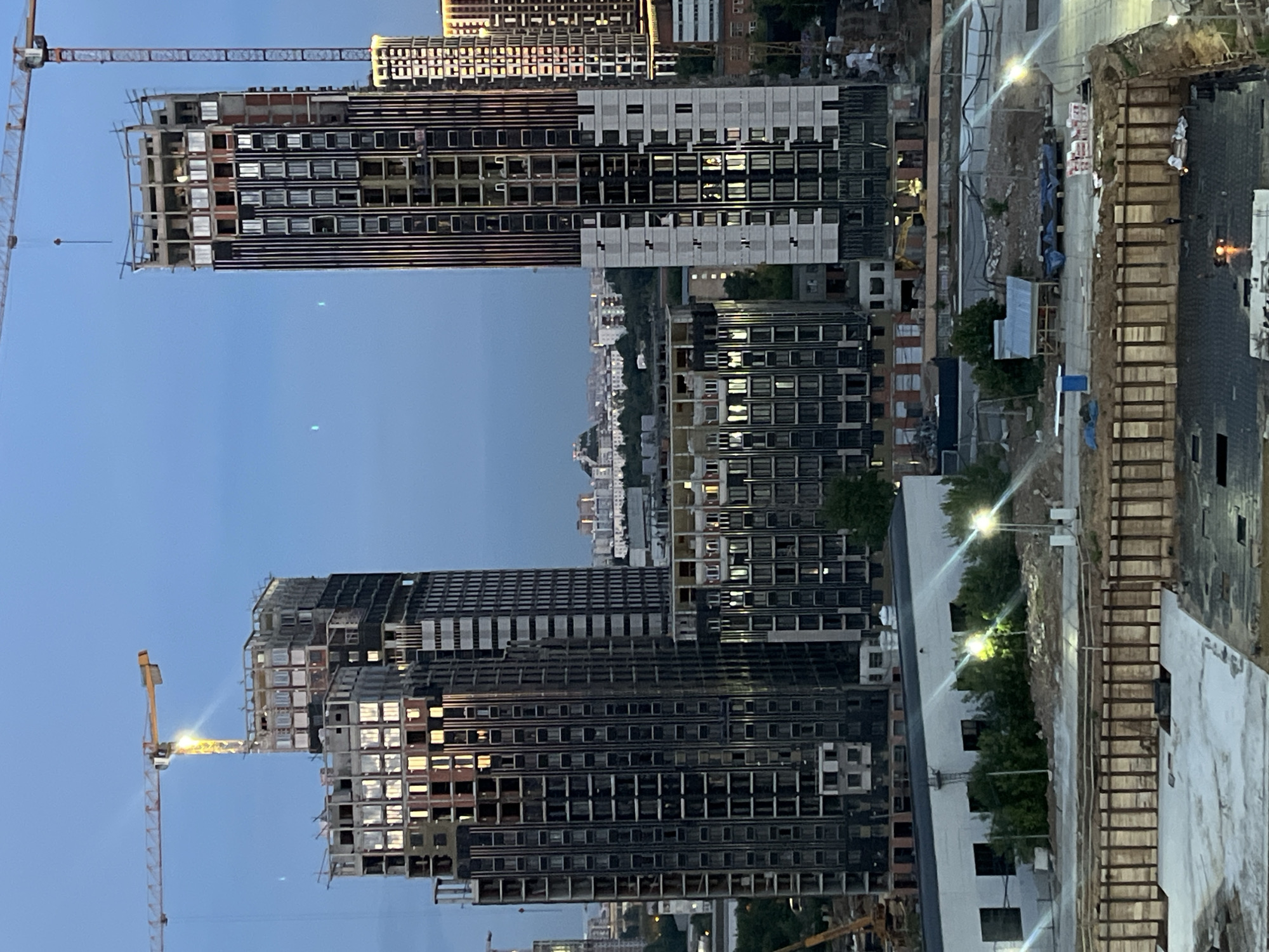 The image captures an aerial view of a construction site in Tarasovka, Russia, during what appears to be twilight. Multiple high-rise buildings are under construction, with their skeletal frameworks and unfinished facades clearly visible. Cranes, essential construction equipment, are prominently featured, one yellow crane with its boom extended over a building, and another red crane to the left of the frame. The scene is dominated by the imposing structures of the buildings, some with dark, reflective windows already installed, while others are still open to the elements.
The time of day is indicated by the soft, diffused light and the illumination from streetlights and lights within the partially constructed buildings, suggesting dusk or early evening. The sky is a clear, pale blue. There are no discernible people or specific weather conditions mentioned. The overall impression is one of active development and urban expansion. The composition emphasizes the scale of the construction project and the transition from day to night.