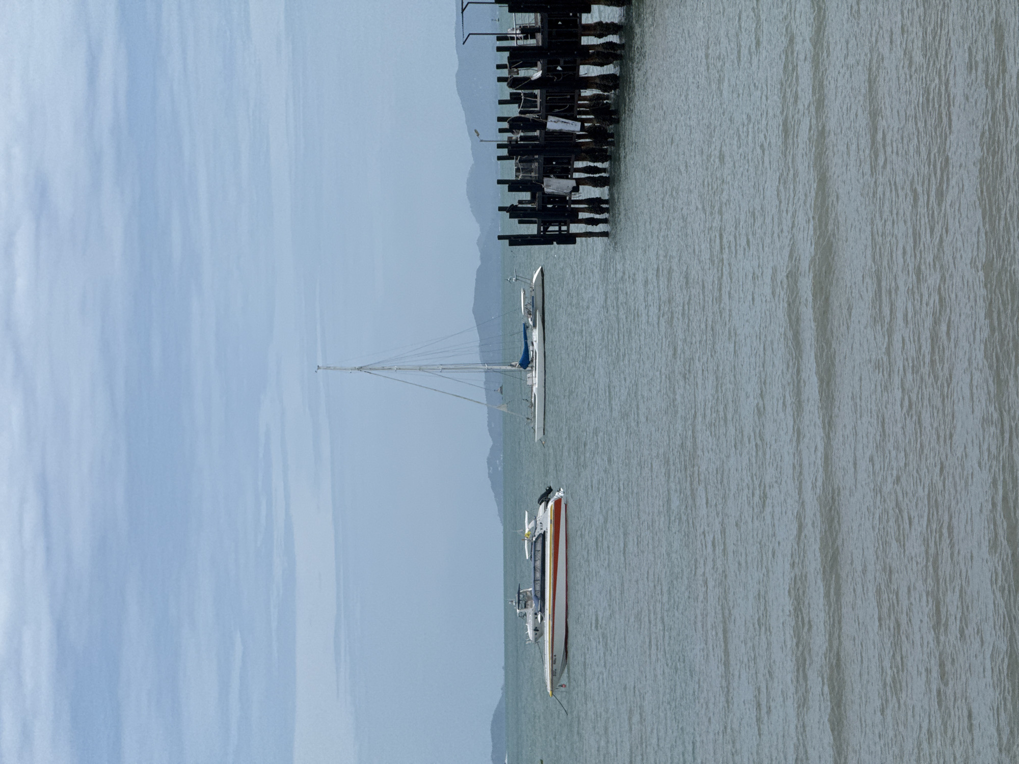 A wide shot shows a serene, overcast day at sea in Ko Samui District, Thailand. The water is a muted grey-green, with gentle ripples disturbing its surface. On the left, a white and orange speed boat is moored, its sleek design suggesting speed. Further back, a large white catamaran with a tall mast and blue sail is anchored, its silhouette sharp against the hazy horizon. In the foreground on the right, a dark wooden pier juts out into the water. The pier is constructed of sturdy, weathered posts, with some white and grey cloths hanging from its structure. In the distance, a chain of lush green islands forms a rugged backdrop, their peaks softened by the atmospheric haze. The sky is a pale blue, streaked with wispy grey clouds, contributing to the tranquil and slightly melancholic mood of the scene.