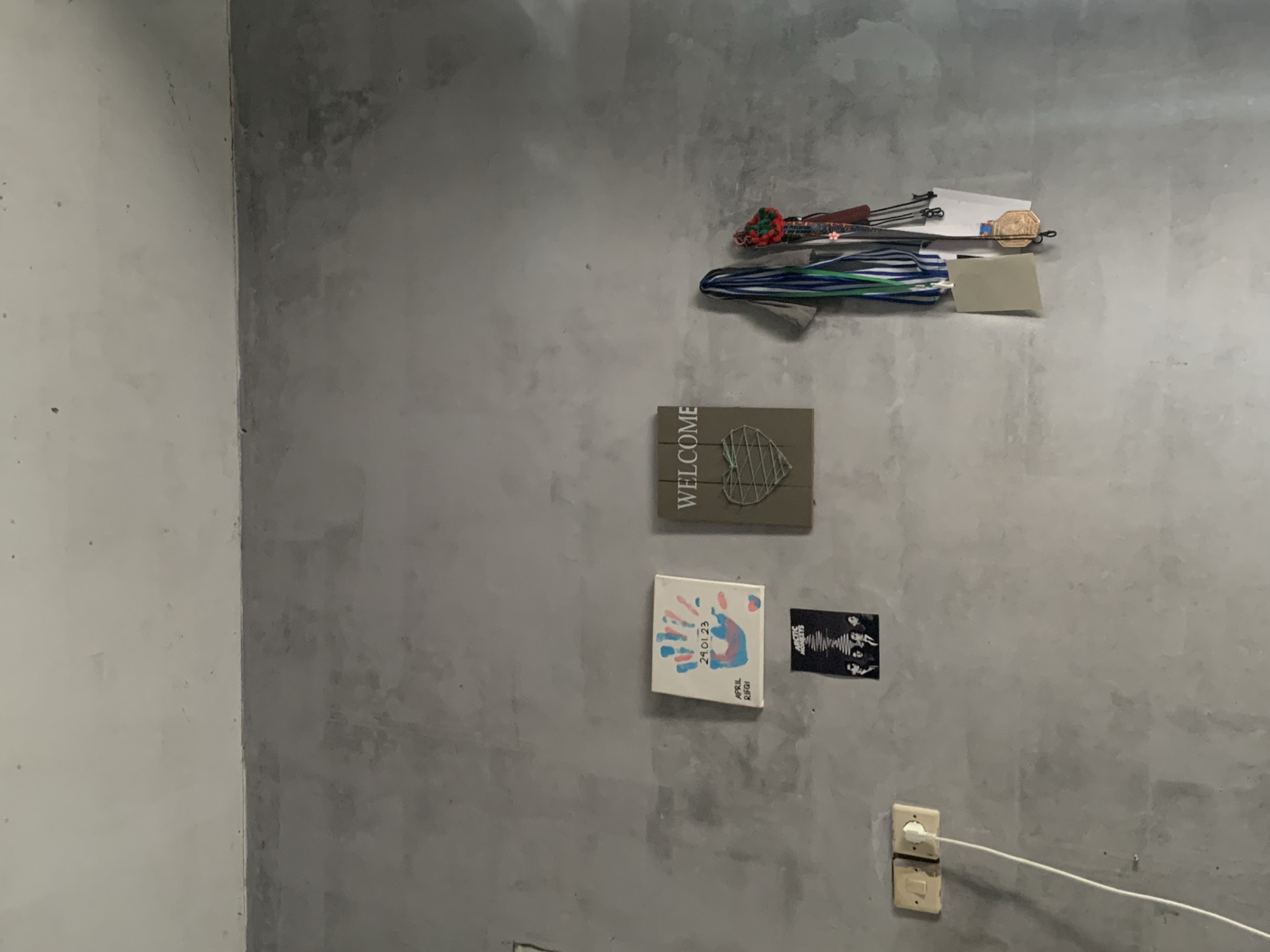 This image depicts a plain gray concrete wall in what appears to be a residential or storage area. On the wall, there are several decorative items and functional fixtures. To the left, there are two light switches, with one connected to a white electrical cord that dangles downwards. Above the switches, a small black poster with the text "ARCTIC MONKEYS" and an image of the band is affixed to the wall. To the right of the poster, a square white canvas displays colorful handprints and the text "24.01.23 APRIL RIFGI". Next to this canvas, a larger wooden plaque with the word "WELCOME" is mounted, featuring a green heart shape made of twine. To the right of the plaque, several lanyards with various colored ribbons and what appear to be medals or awards are hanging from hooks. The overall environment is simple and sparsely decorated, suggesting a utilitarian space.