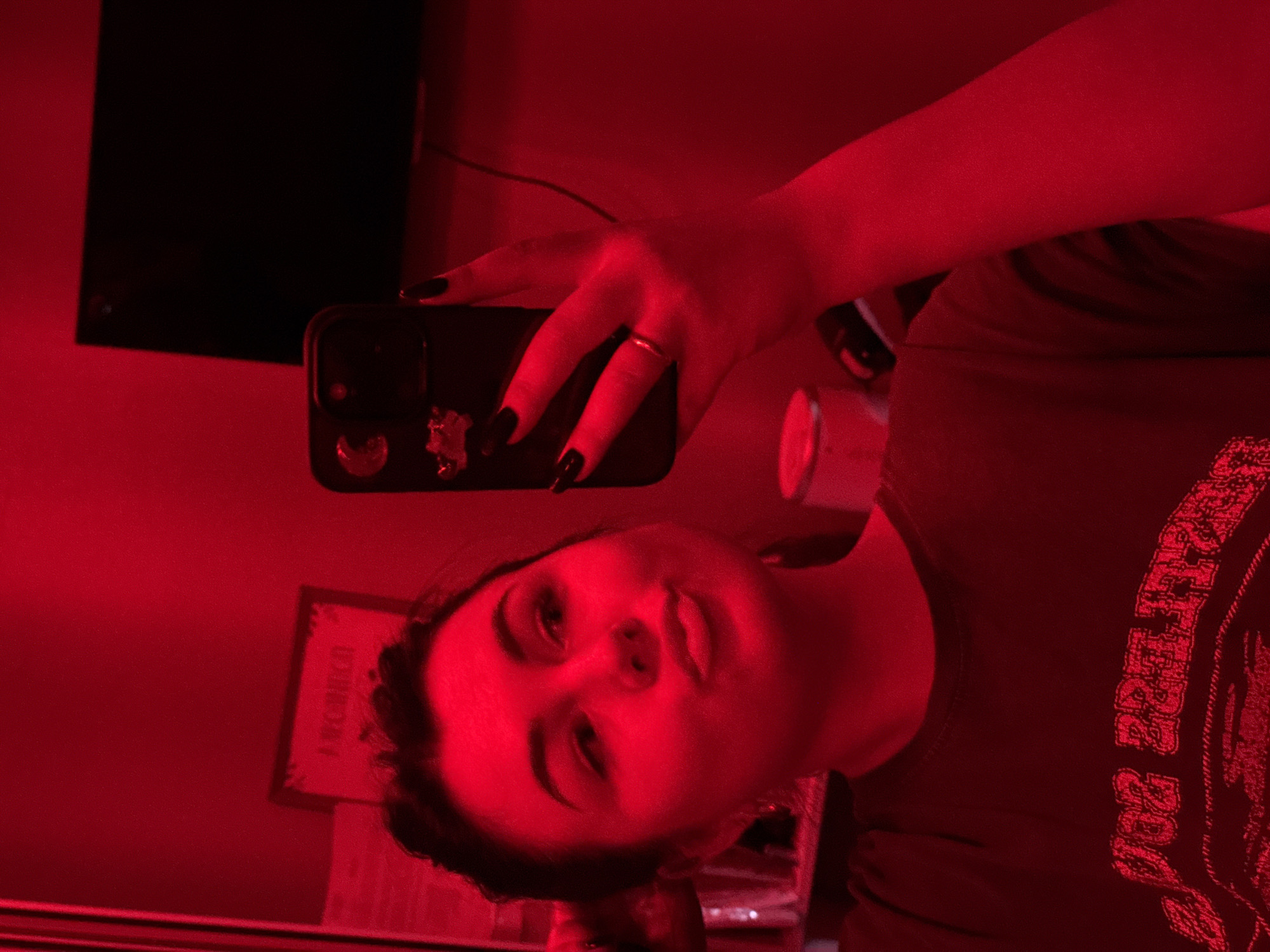 A person with dark hair and a red-toned complexion, likely due to the lighting, is taking a selfie with a black smartphone. They are wearing a ring on one finger. The environment is dominated by red light, suggesting a red light district. A black, rectangular object, possibly a television screen, is visible in the background. An item with illegible writing on it is partially visible on the wall behind the person. The city of Chaikovsky, Russia is not determinable from this image.