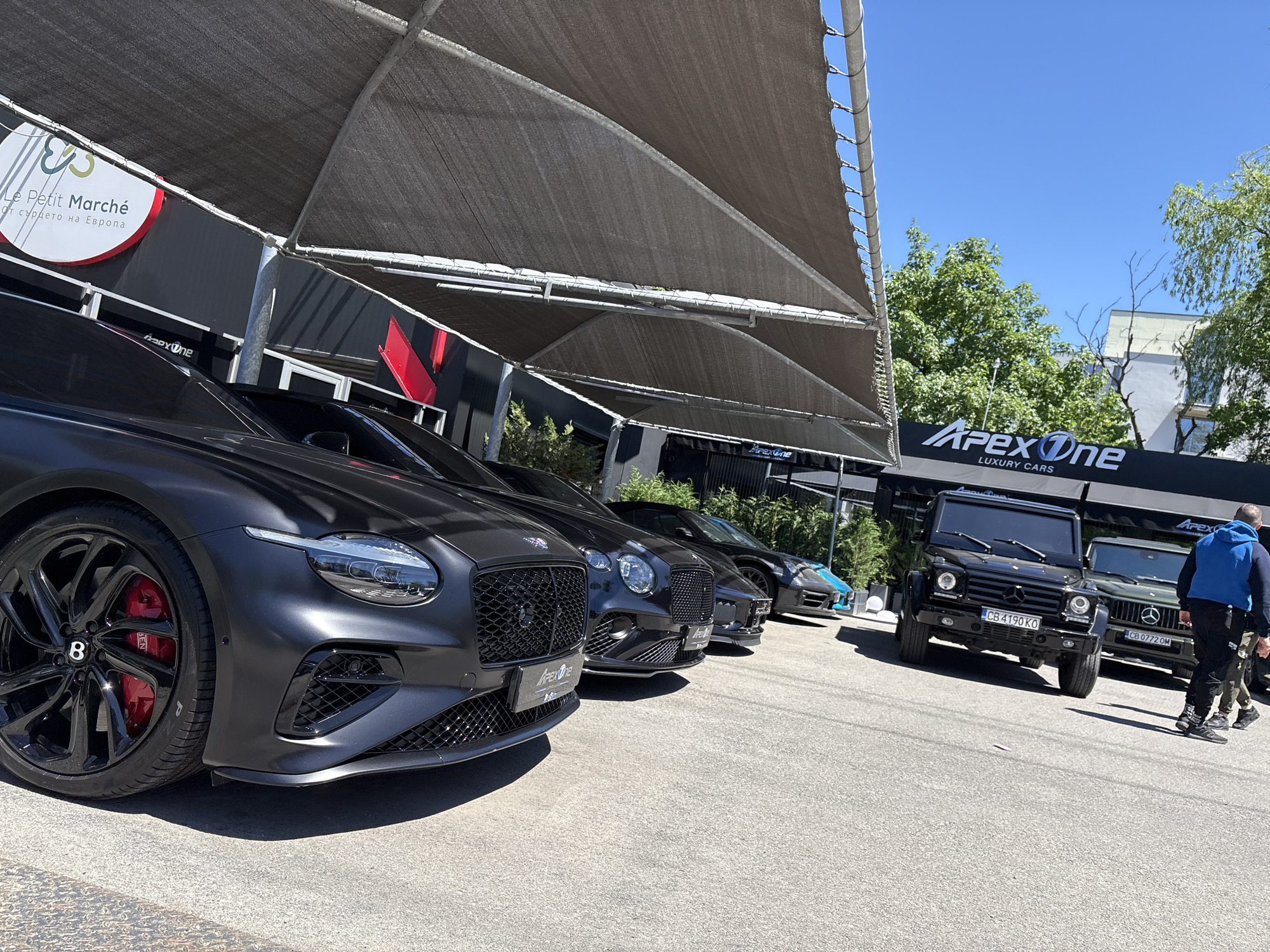 The image depicts a luxury car dealership, "Apex One Luxury Cars," in Sofia, Bulgaria. Several high-end vehicles, including Bentleys and Mercedes-Benz G-Wagons, are parked under a canopy outside the dealership. A sign for "Le Petit Marché - От сърцето на Европа" is also visible on the building. A man wearing a blue hoodie stands near the G-Wagons on the right side of the frame. The sky is clear and blue. Vehicle license plates are visible: "CB 4190 KO" and "CB 0772 OM."