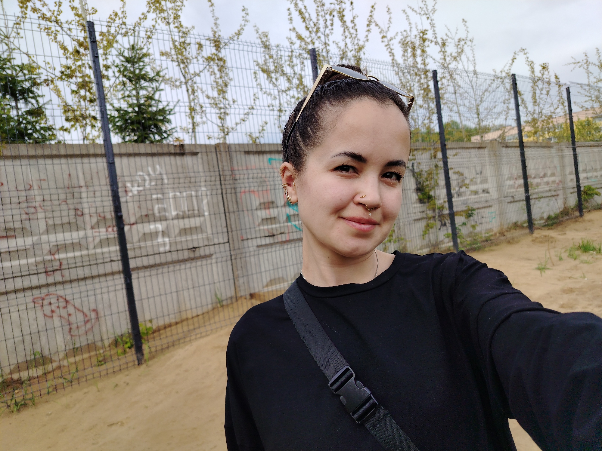 A medium, eye-level shot captures a woman posing for a selfie in front of a concrete and metal fence. The woman has her hair pulled back, and sunglasses are perched atop her head. She wears a black top and a strap across her chest. Her face is turned slightly to the right, and she smiles. Several piercings adorn her ear and nose.
Behind her, a concrete wall runs horizontally across the frame, topped with a wire fence. Trees are visible behind the fence, partially obscuring the sky. The ground appears to be a sandy or dirt surface. Based on visual cues, the location may be near a residential or urban area. The location of Noviy, Russia is assumed.