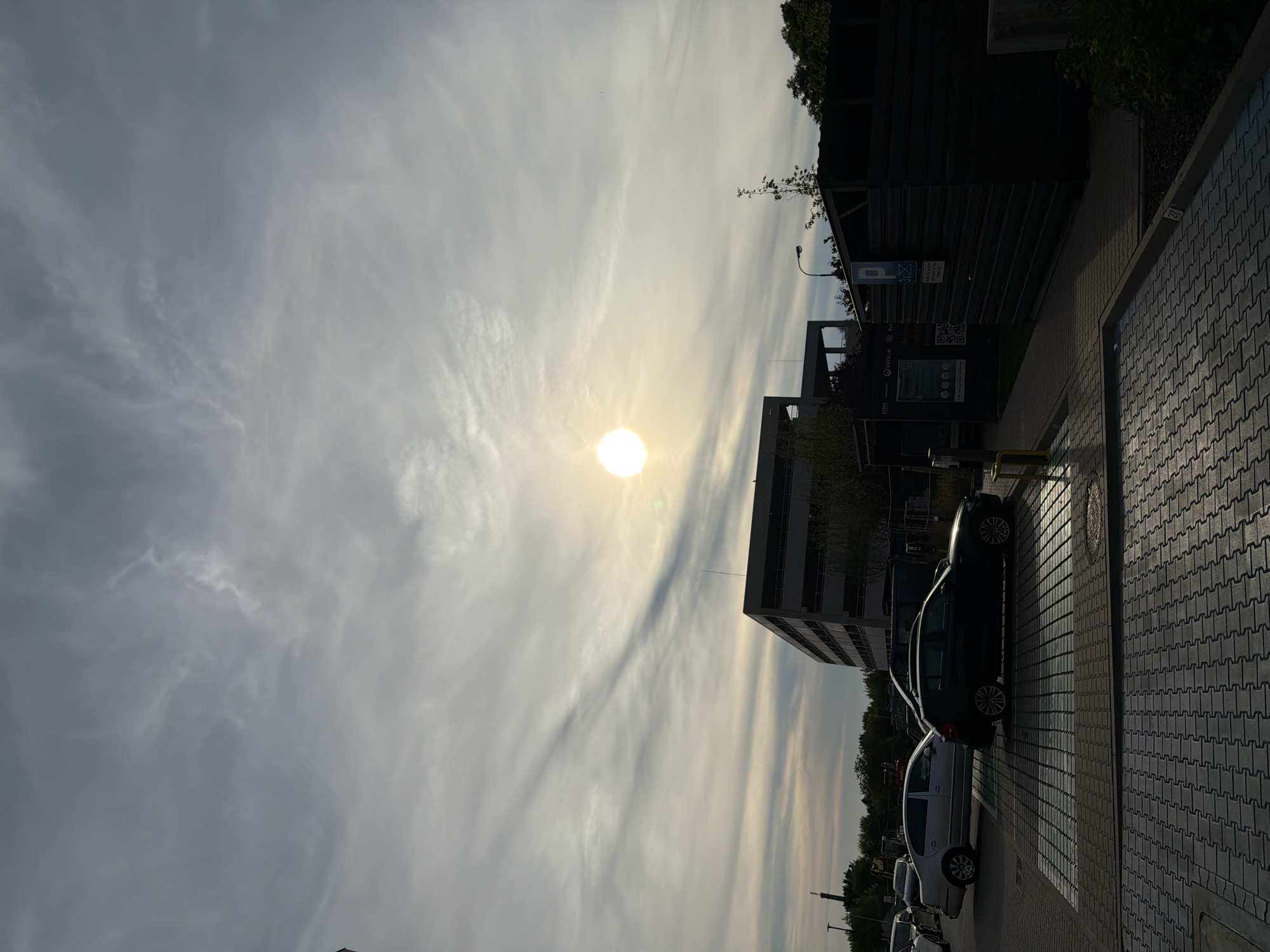 The image depicts a low-angle view of a parking lot with several cars, a multi-story building, and a dark, corrugated metal structure under a sky filled with wispy clouds and a bright, low sun. The sun is visible in the upper center of the frame, casting a glare. The parking lot is paved with interlocking bricks. In the foreground, there are several parking spaces. A silver sedan is parked on the left, and a dark hatchback is in the center. Behind them, a large, dark structure with a pitched roof is partially visible. To the right, a dark, utilitarian building with horizontal corrugated metal siding is prominent. A blue parking sign with a white "P" and a number is affixed to its side. The multi-story building in the background is grey and has multiple levels of open-air parking or balconies. The scene appears to be an outdoor commercial or office complex. The lighting suggests either sunrise or sunset. This image is likely from the immediate vicinity of the Łódź University of Technology, specifically near the Faculty of Mechanical Engineering, given the architectural style of the buildings and the presence of a parking area typical for such institutions in Łódź, Poland.