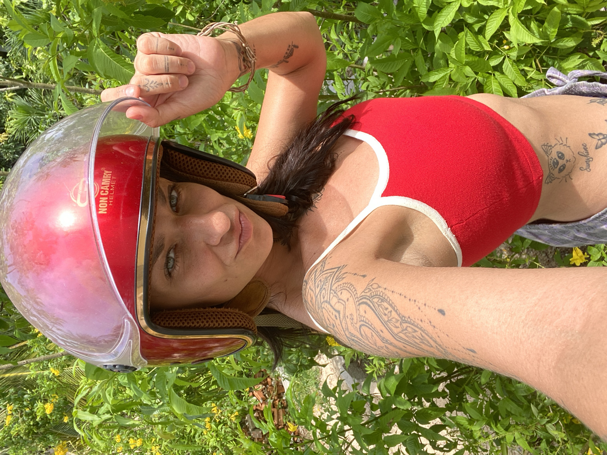 The media file features a woman wearing a red top and grey shorts, posing outdoors with a red helmet. She has several tattoos visible on her arm and torso, including a whimsical drawing of a creature on her abdomen. The setting appears to be a tropical environment, with lush green foliage and yellow flowers surrounding her. The lighting suggests it is daytime, likely sunny, casting shadows and highlighting the textures of the leaves and the woman's skin. The woman's gaze is directed towards the camera, with a neutral or slightly curious expression. Text on the helmet reads "NON CAMRY HELMET". The overall mood is casual and relaxed, possibly taken during a break from travel or leisure activities in a warm, sunny climate like Mũi Né, Vietnam.