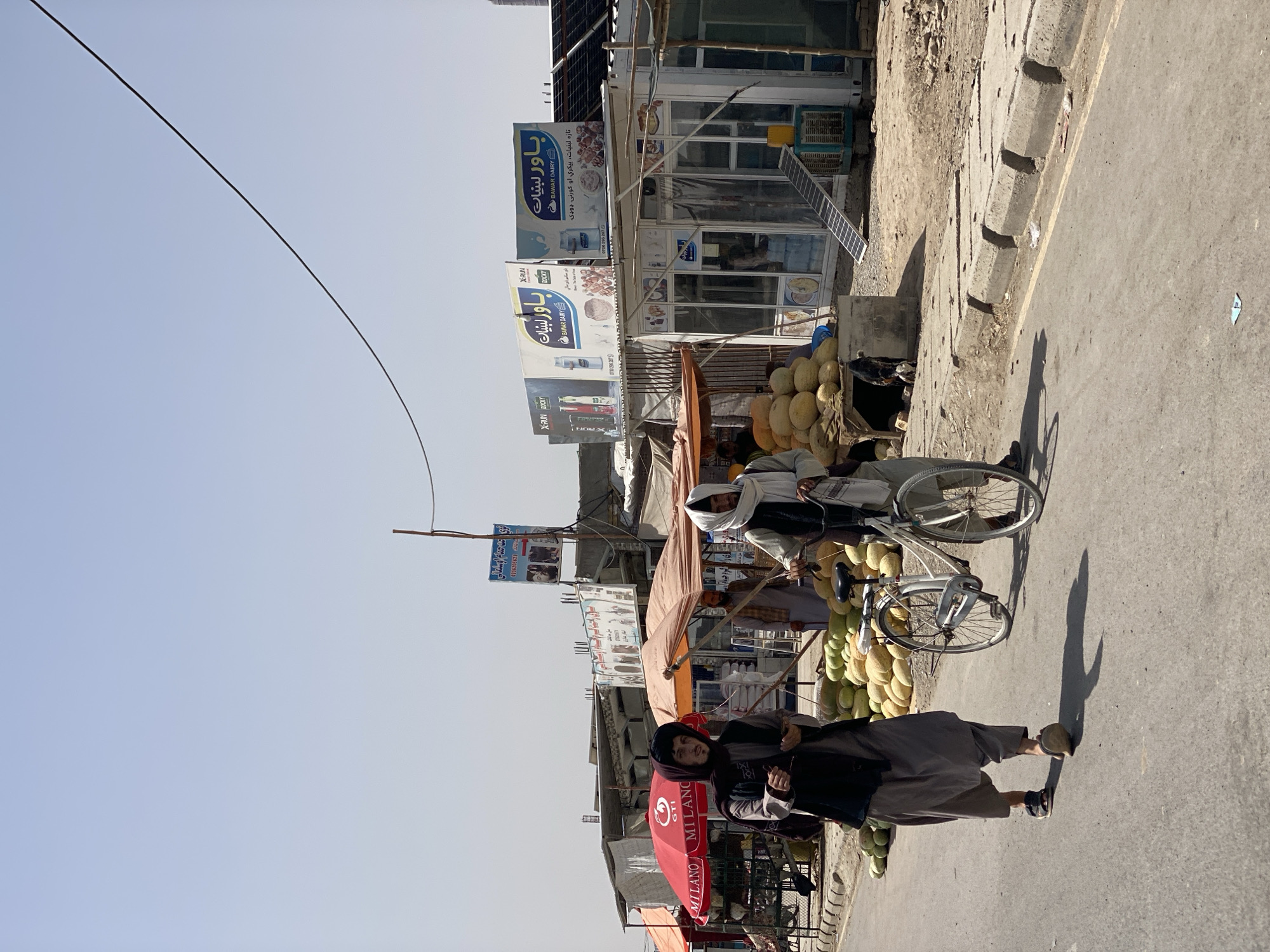 In Kandahar, Afghanistan, two individuals are walking on a paved street next to a vendor stall laden with melons. A white bicycle is positioned between them, with one person leading it. The vendor stall features a wooden stand with a canopy and more melons, suggesting a marketplace or roadside produce stand. Behind the stall, a building displays advertisements for "BAWAR DAIRY." The environment appears to be a bustling urban setting with shops and signs indicating local commerce. The sky is clear and bright.