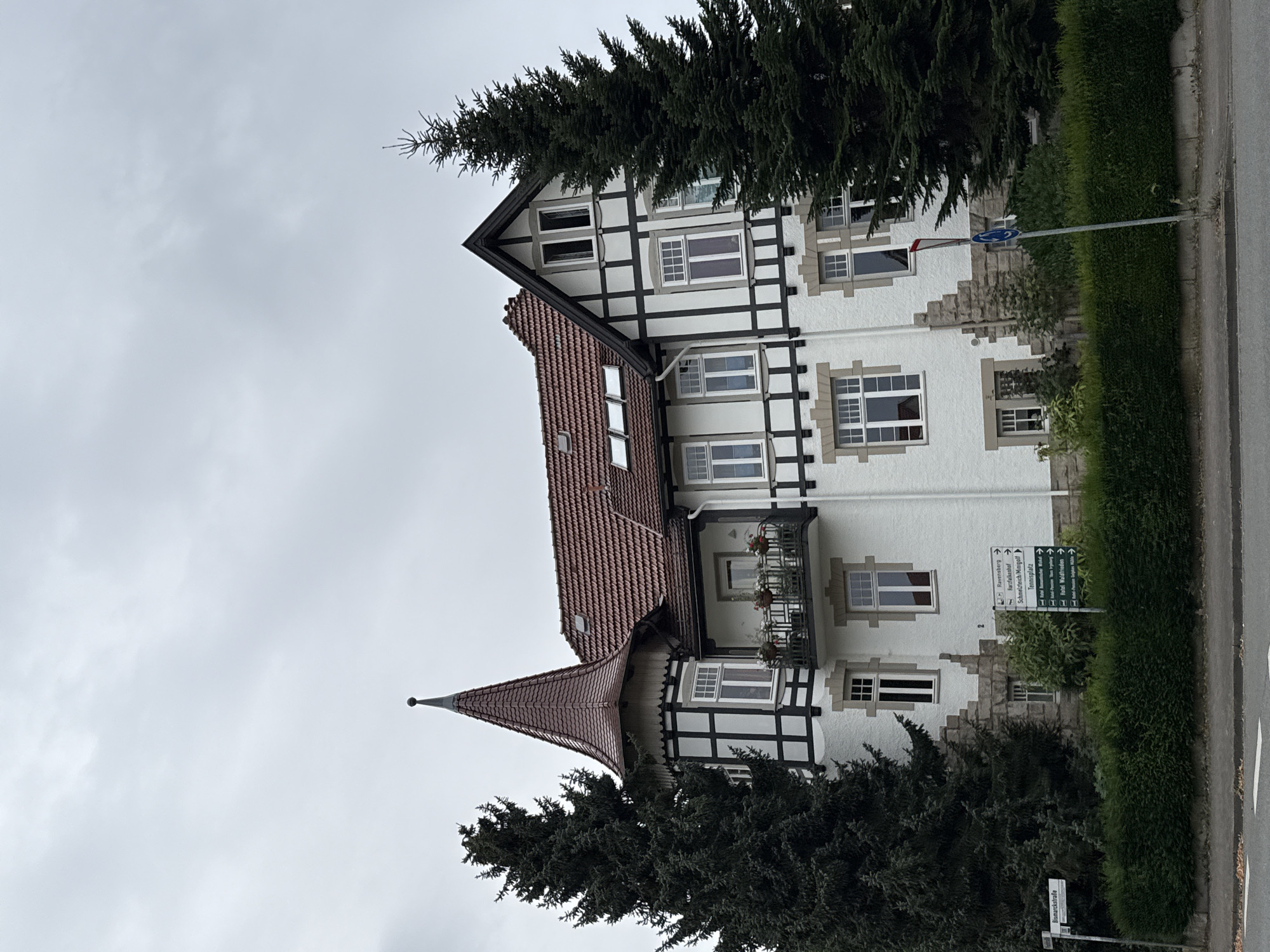 The image shows a large, white building with a dark, timber-framed upper story and a red-tiled roof. The building has several windows, a balcony, and a prominent conical turret with a spire. Two large evergreen trees flank the building. In front of the building, there is a dense green hedge. A paved road is visible in the foreground. A street sign in German that reads "Bismarckstraße" is to the left of the building. To the right of the building, a directional signpost with several green signs pointing to various locations, including "Ravensberg", "Karzafalkenhof", and "Hotel Waldrieden," is visible. The sky is overcast and gray. The scene depicts a building in Bad Sachsa, Germany.