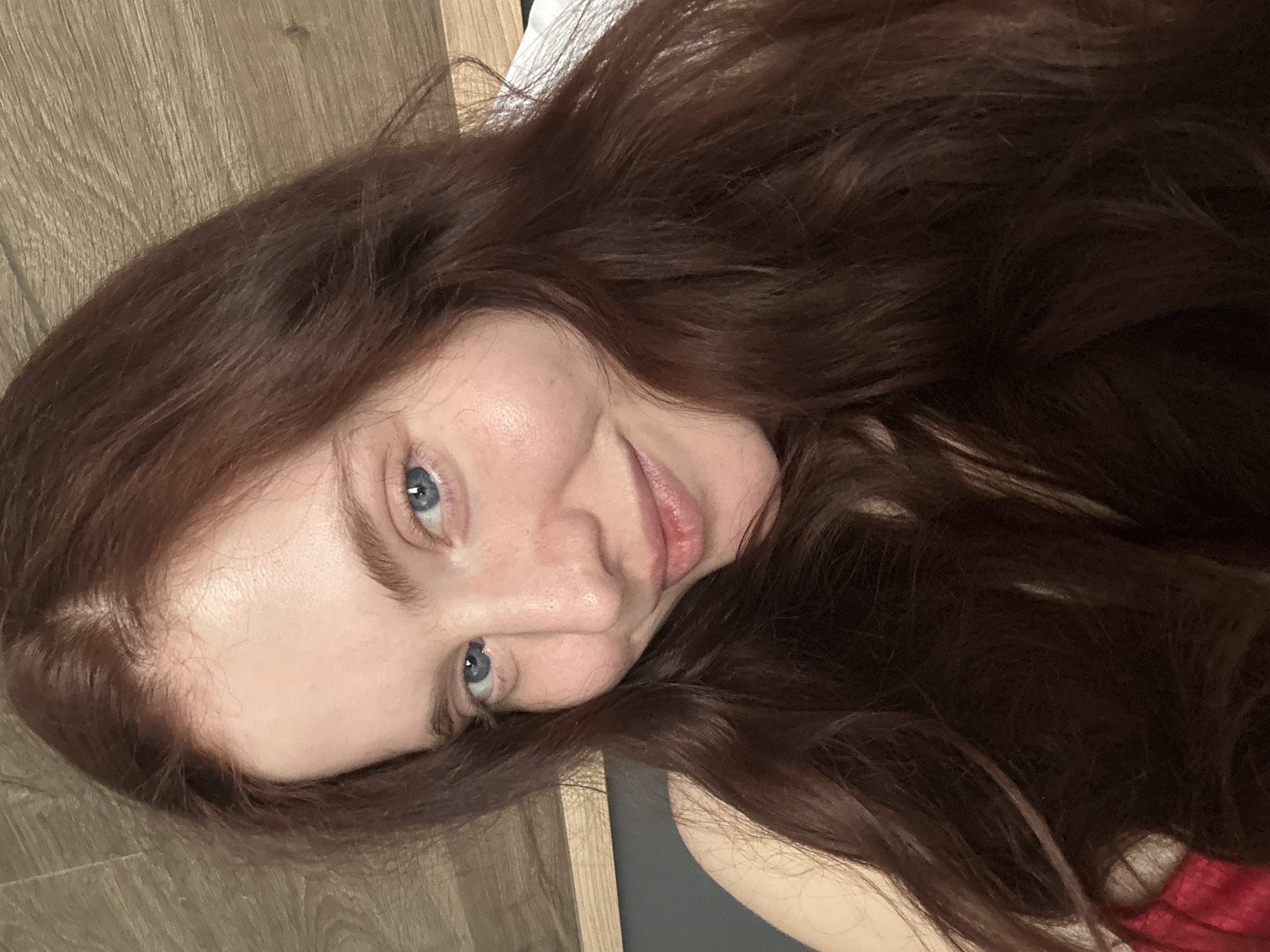 A woman with long, wavy, auburn hair and blue eyes is shown in a close-up, slightly angled portrait. The background includes a wood-grain surface (possibly a floor) and a neutral-toned wall or object, suggesting an interior setting. The woman's expression is somewhat playful, with a slight smile. There's no specific indication of Riga, Latvia, in the image.