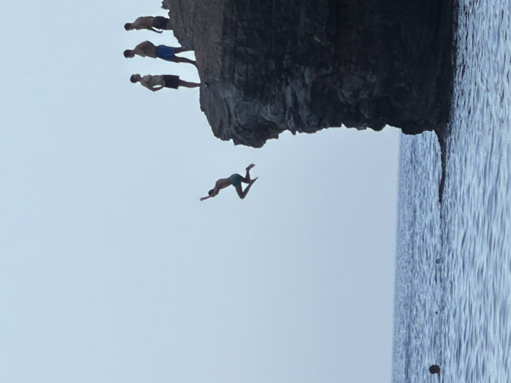 A group of young men are cliff diving into the Adriatic Sea off a rocky outcrop on the coast of Budva, Montenegro. One man is captured mid-air, mid-jump, having launched himself from the cliff face. Several other men stand on the cliff edge, some watching the diver, others preparing to jump. The sea is calm with gentle waves, and the sky is a clear, pale blue. The scene evokes a sense of adventure and exhilaration.