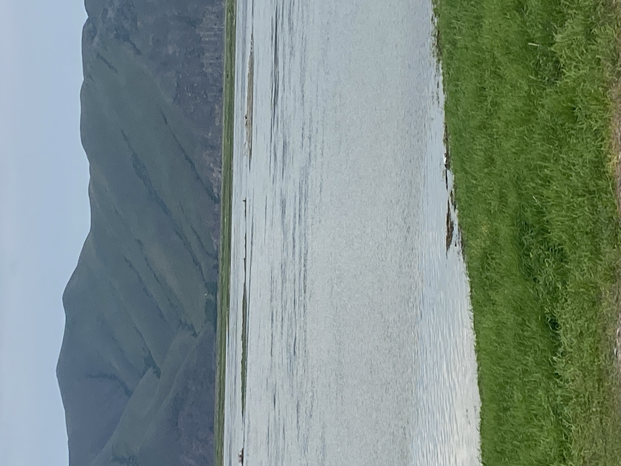 The image shows a wide body of water, likely a river or lake, with grassy shores. In the background, rolling green hills form a natural landscape. The sky is overcast, suggesting a cloudy day. There are two small boats visible on the water, one further in the distance and another closer to the left side of the frame. A patch of land or a small island is present in the middle of the water. The foreground is dominated by lush green grass. The image depicts a natural scenic view near Chernogorsk, Russia.