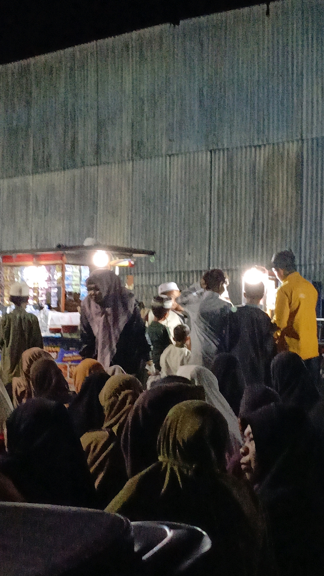 The image depicts an outdoor gathering at night in Kota Pagatan, Indonesia. A group of individuals, some wearing traditional Islamic attire such as kufis and hijabs, are congregated in front of a makeshift stall illuminated by bright lights. The stall appears to be selling items displayed on shelves and appears to be a food or goods vendor. In the background, a corrugated metal wall serves as a backdrop. The lighting suggests it is nighttime, and the overall atmosphere seems to be one of community interaction or a market event. Some people are looking towards the stall, while others are in conversation with each other. The ground is not clearly visible, but the scene appears to be taking place on a level surface.