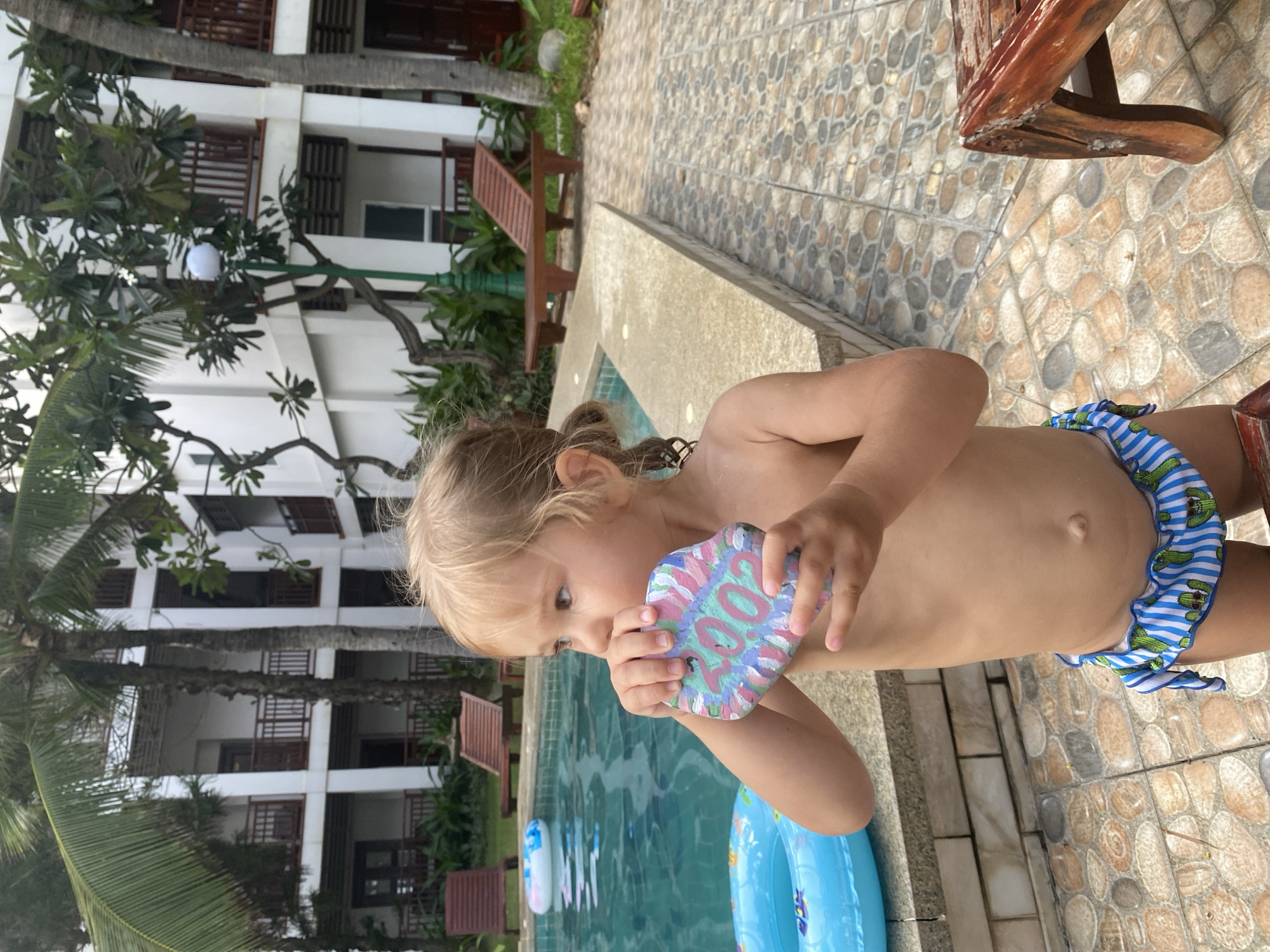 A young child, likely a girl due to her blonde hair and bathing suit, stands by the edge of a swimming pool. She is holding a decorated rock, which appears to have a colorful, abstract design on it. The child is wearing a blue and white striped bathing suit with a ruffled waistband adorned with small cactus patterns. The swimming pool water is a clear blue, with two inflatable rings floating on its surface. The poolside is paved with decorative stone tiles.
In the background, a multi-story building with balconies and wooden shutters is visible, surrounded by lush green palm trees and other tropical foliage. There are also wooden lounge chairs placed on the poolside. The lighting suggests it is daytime, with diffused sunlight. The scene evokes a relaxed, vacation-like atmosphere, possibly at a resort or hotel in a tropical location. The child's focus on the painted rock suggests an element of play or creative engagement. The overall impression is one of a peaceful, summery day. The text "2022" is visible in white paint on the decorated rock. The location is Phu Thuy Ward, Vietnam.