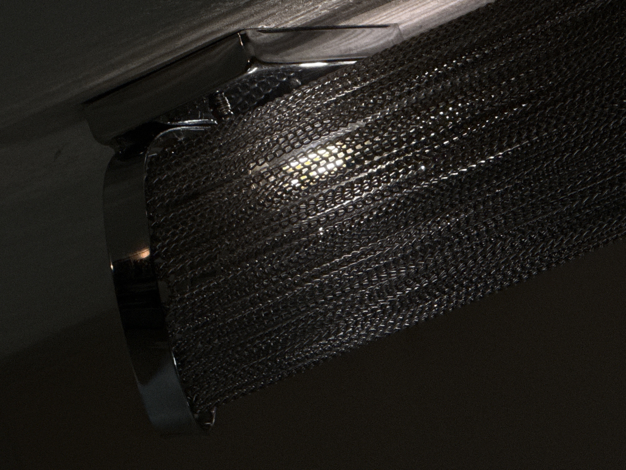 This is a close-up, low-angle shot of a showerhead and a stream of chain-like material, possibly from a decorative shower curtain, flowing downwards in a dimly lit environment. The showerhead is metallic and appears to be a modern design. The chain material is dense, forming a curtain-like effect, with light reflecting off its links. The background is dark, with some textured surface visible on the right side, possibly a wall. There is no indication of any specific location in Yuzhno-Sakhalinsk, Russia, or any people or actions taking place. The scene appears to be staged or an artistic representation.