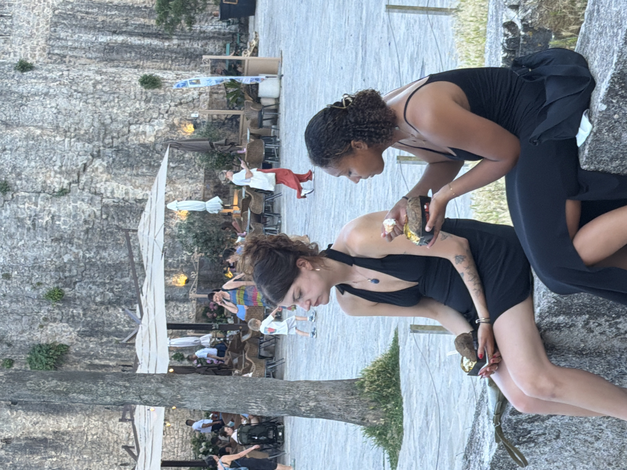 Two young women are sitting on a stone wall, eating from take-away containers filled with food. The woman on the left has dark hair pulled up, a black halter dress, and is holding her container. She has tattoos on her left arm and is wearing a necklace with a blue pendant. The woman on the right has curly dark hair, a black spaghetti strap dress, and is holding her container. She is wearing a necklace with a thin chain. They are outdoors in front of a large, rough stone wall. In the background, there are outdoor seating areas with umbrellas and tables, suggesting a restaurant or cafe. People are visible in the background, some walking and some seated. The scene appears to be in Budva, Montenegro, likely near the Old Town fortifications.