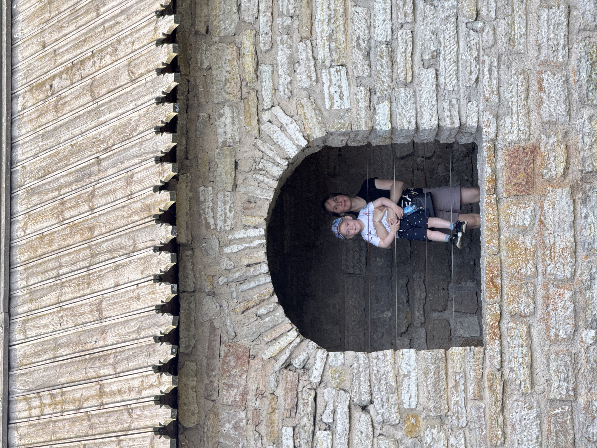 A medium shot captures a woman holding a young child in front of a rough-hewn stone archway of a building that appears to be part of the Oreshek Fortress in Shlisselburg, Russia. The woman, wearing a black t-shirt, stands to the right of the frame, with the child facing forward in her arms. The child is wearing a white t-shirt, dark shorts with a patterned design, a baseball cap, and dark sneakers. The stone wall is constructed from irregular blocks of varying sizes and colors, with visible mortar. Above the archway, a weathered wooden roof with vertical planks is visible, featuring decorative eaves. The background within the archway is dark and indistinct. Two thin wires are stretched horizontally across the archway opening. The lighting suggests an outdoor setting during the daytime.