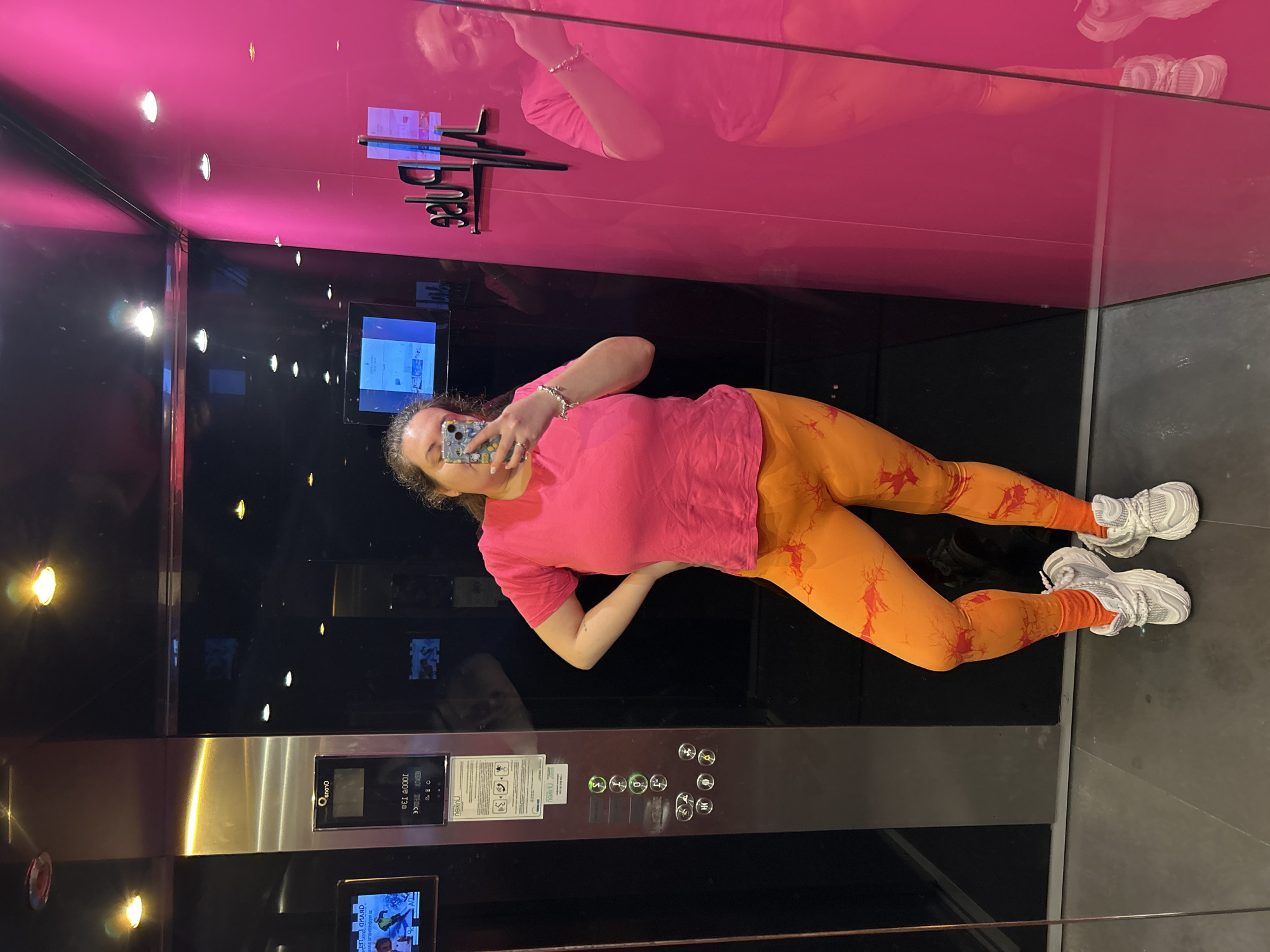 A woman is taking a selfie in an elevator. The elevator has a pink top, black middle and grey bottom, with elevator buttons on the right. The woman is wearing a pink t-shirt, orange leggings, and white sneakers. The location is Plovdiv, Bulgaria.