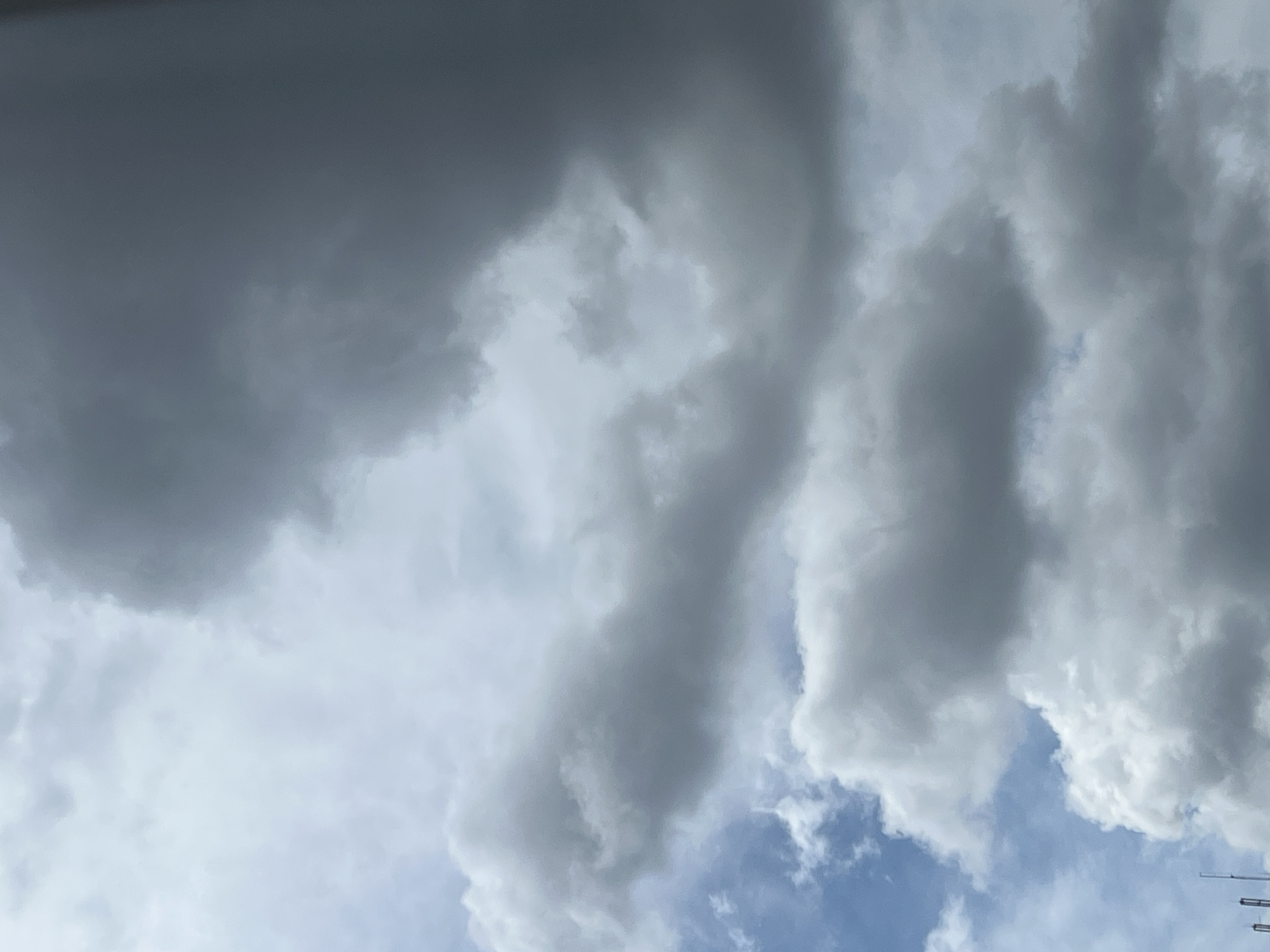 The image shows a sky filled with dense, dark gray clouds, indicative of impending or ongoing rainfall. The lower portion of the frame reveals fragments of blue sky and lighter, fluffier white clouds. In the very bottom left corner, the tips of several tall, thin structures, likely communication towers or construction cranes, are visible. The scene is a wide shot, capturing a dramatic sky over what appears to be an urban environment, though no specific landmarks of Warsaw, Poland, are identifiable.